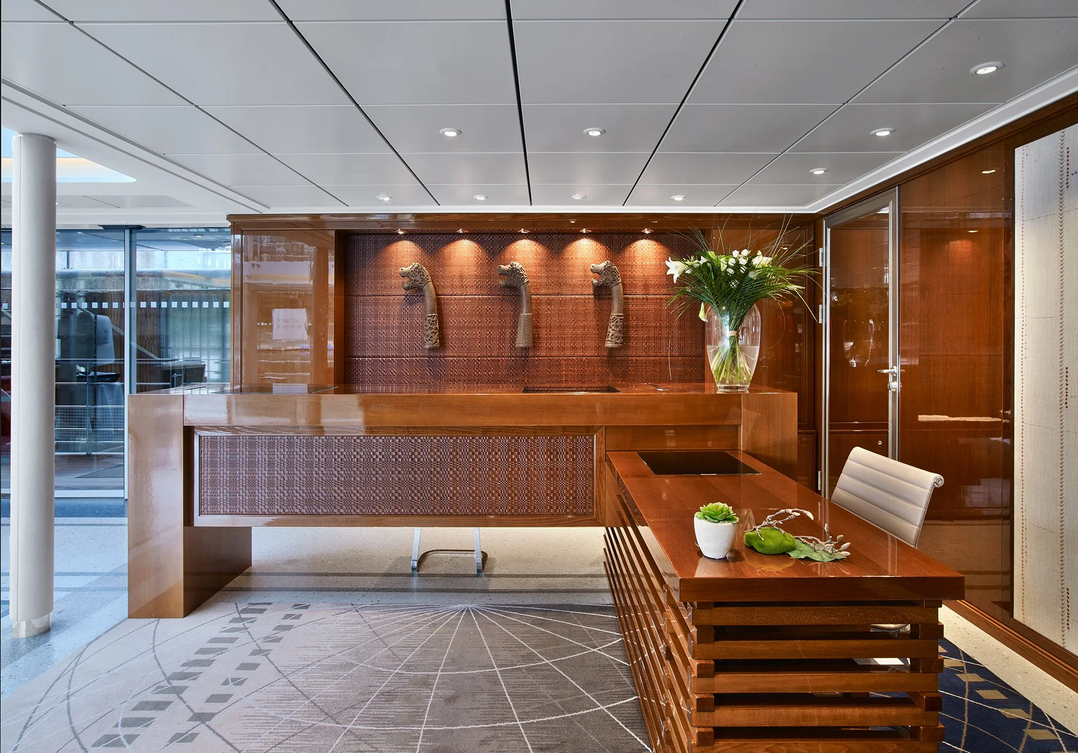 Hotel reception area with a wooden desk, decorative wall with three animal head sculptures, a glass vase with white flowers, a small potted plant, and a cream-colored chair.