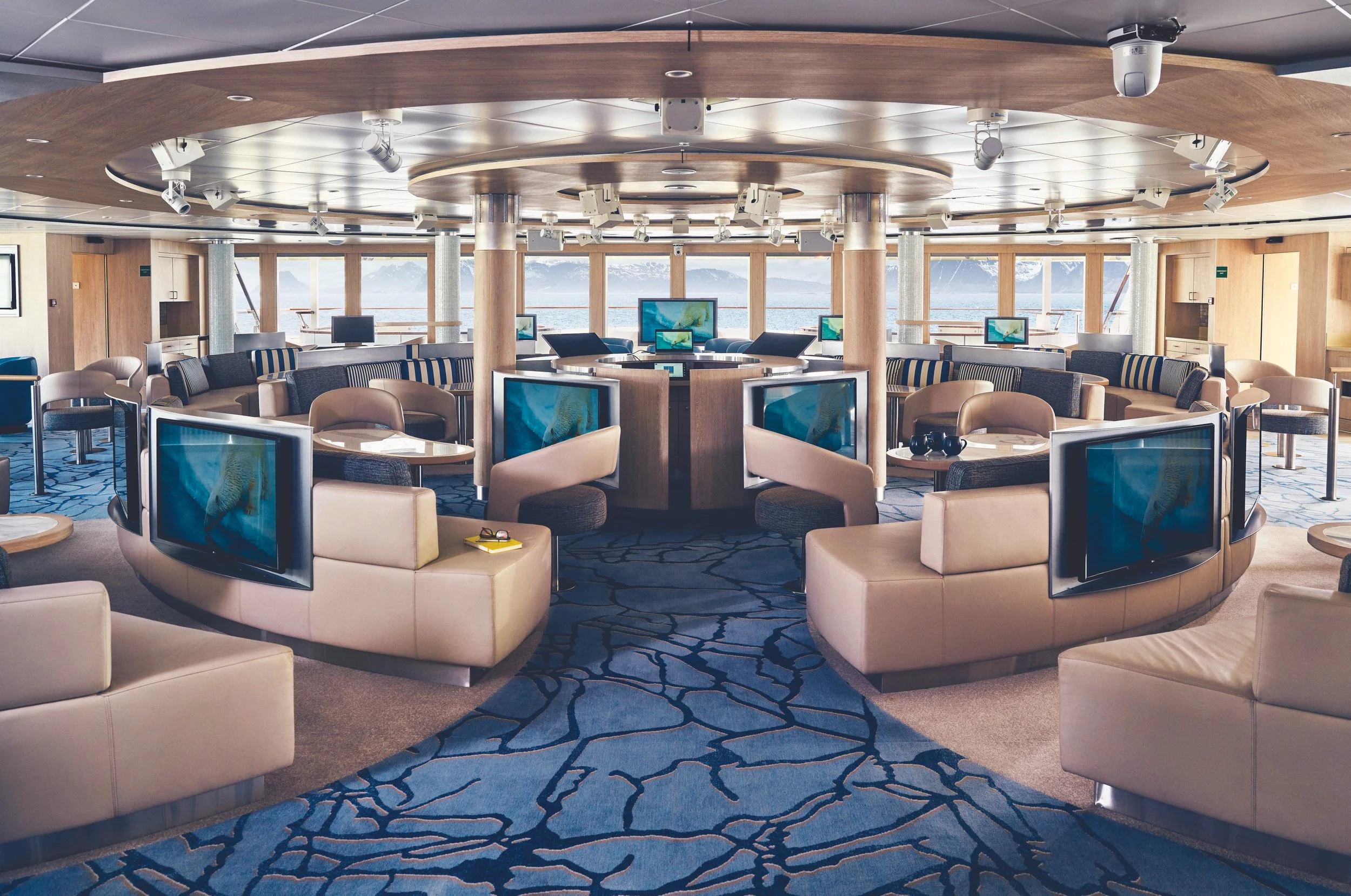 A large, modern lounge area on a cruise ship with multiple flat-screen TVs, beige and dark blue seating, and panoramic windows showing water and mountains in the background.