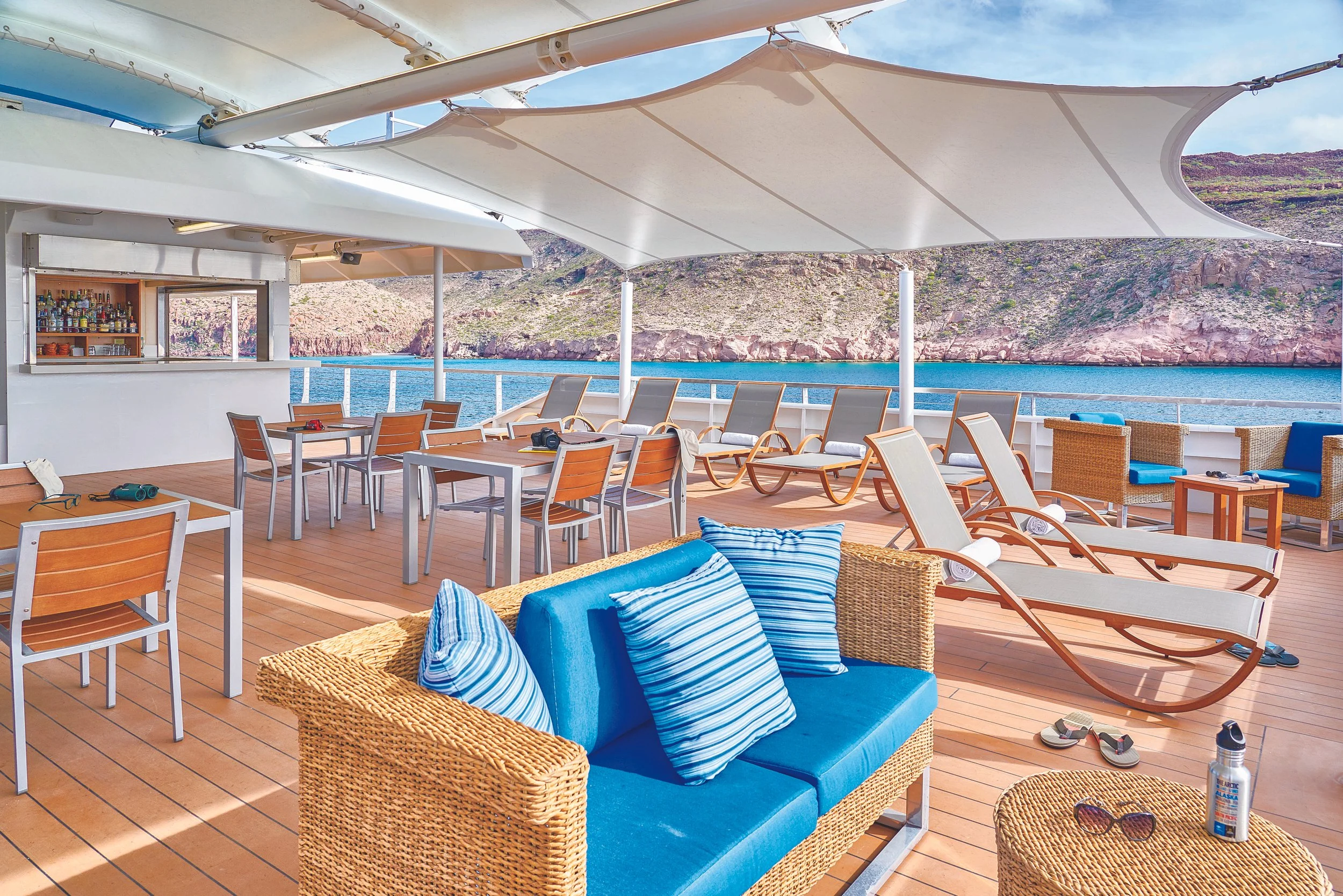 Deck of a cruise ship with lounge chairs, tables, umbrellas, a bar, and outdoor seating, overlooking a body of water with rocky hills in the distance.