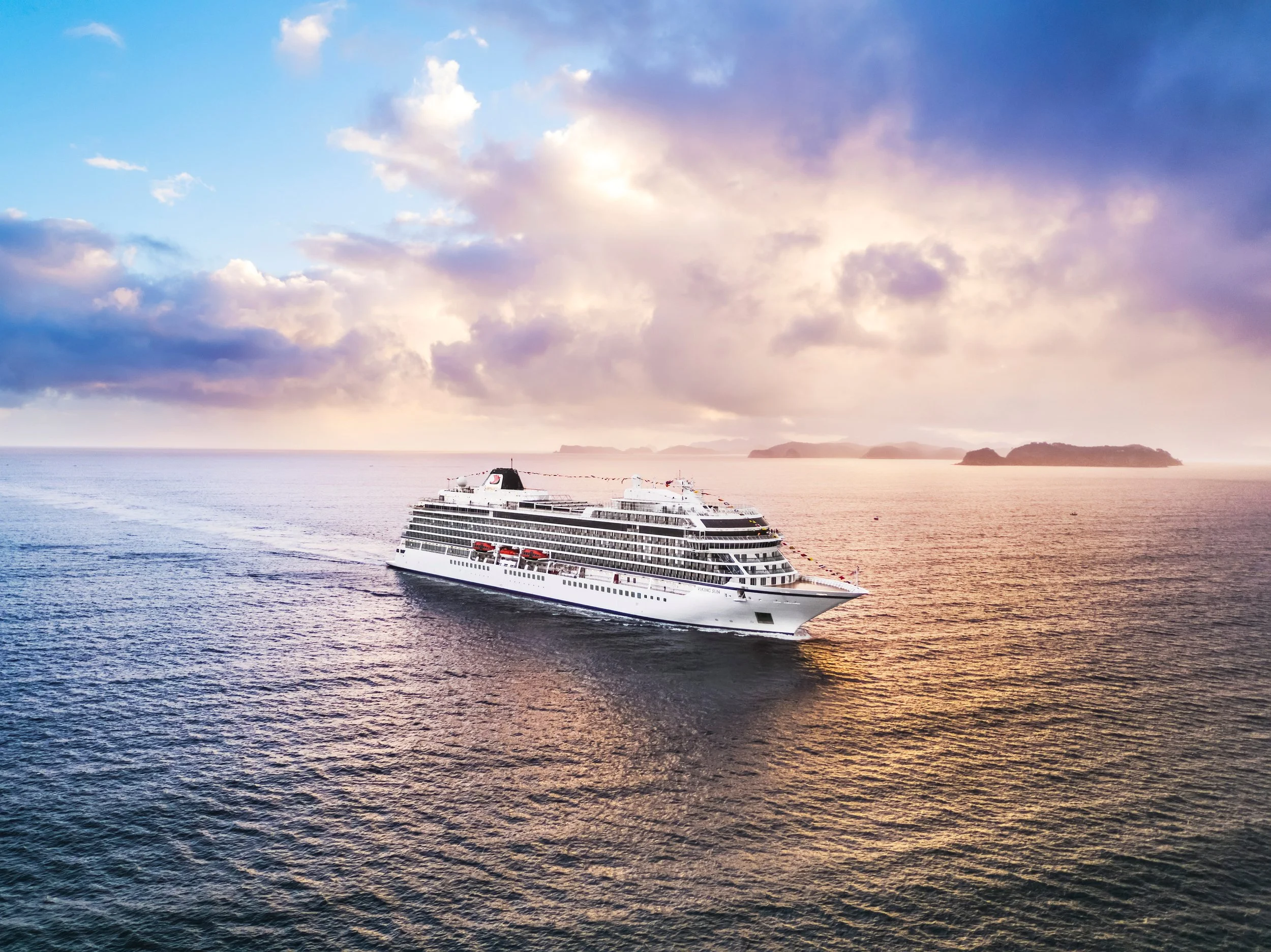 A large white cruise ship sailing on calm ocean waters during sunset, with distant islands and a partly cloudy sky.