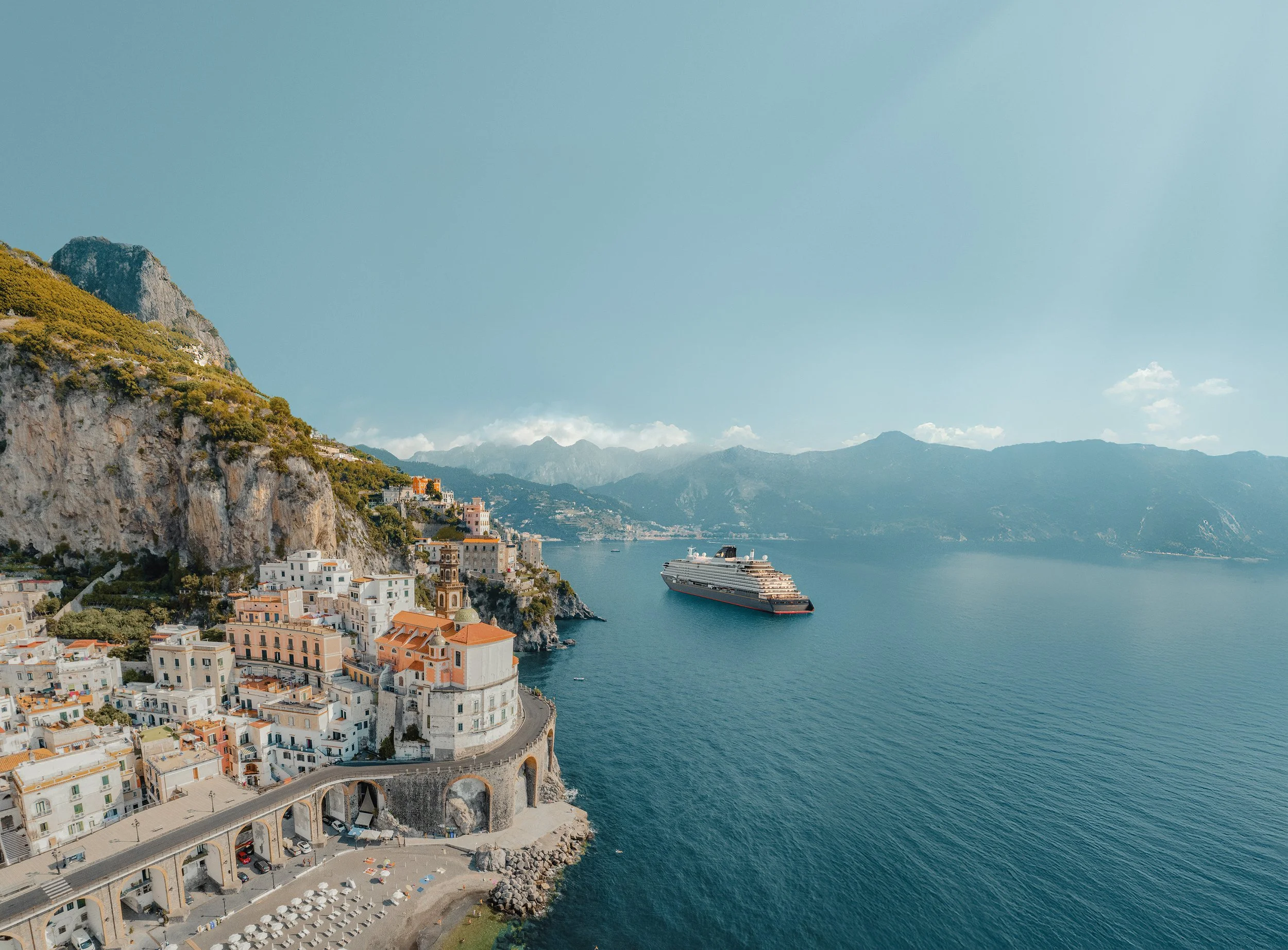 Coastal town with colorful buildings on a hillside, a street with parked cars and umbrellas, a large cruise ship on the calm blue sea, and mountains in the background under a clear sky.