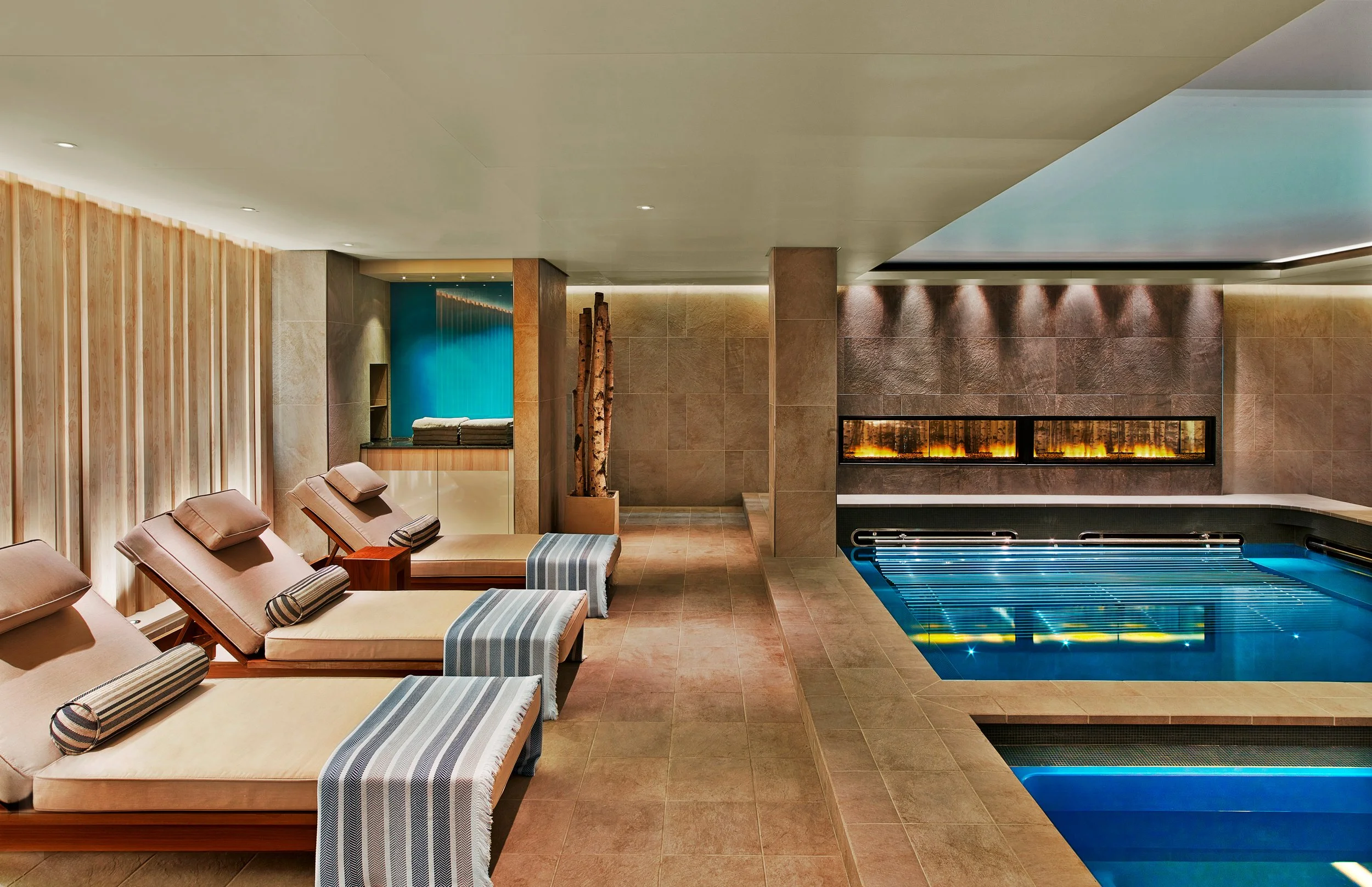 Indoor spa area with four lounge chairs, small side tables, a fireplace, and a swimming pool.