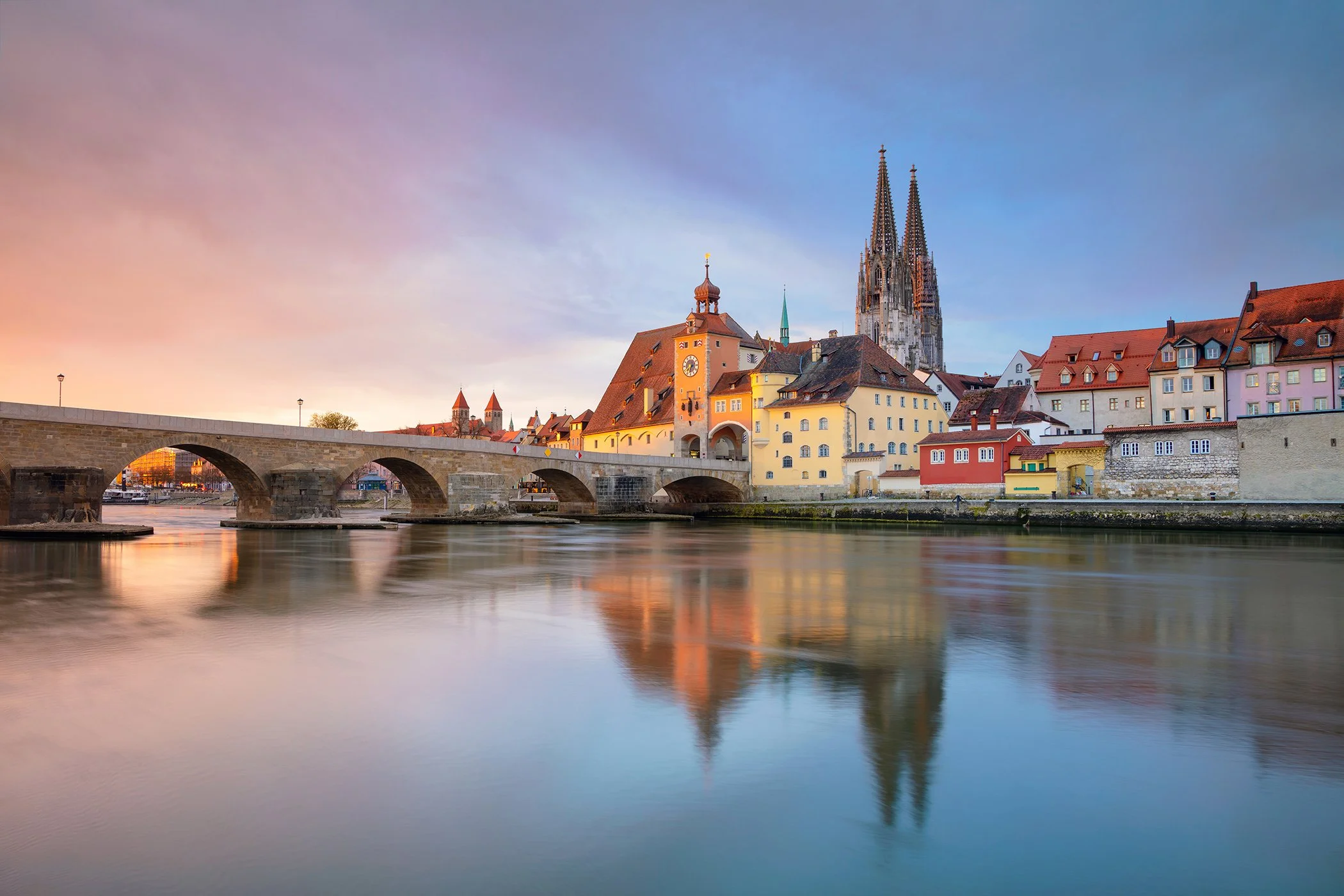 Scenic view of a historic European town with colorful buildings, a church with tall spires, and a stone bridge over a calm river during sunset or sunrise.