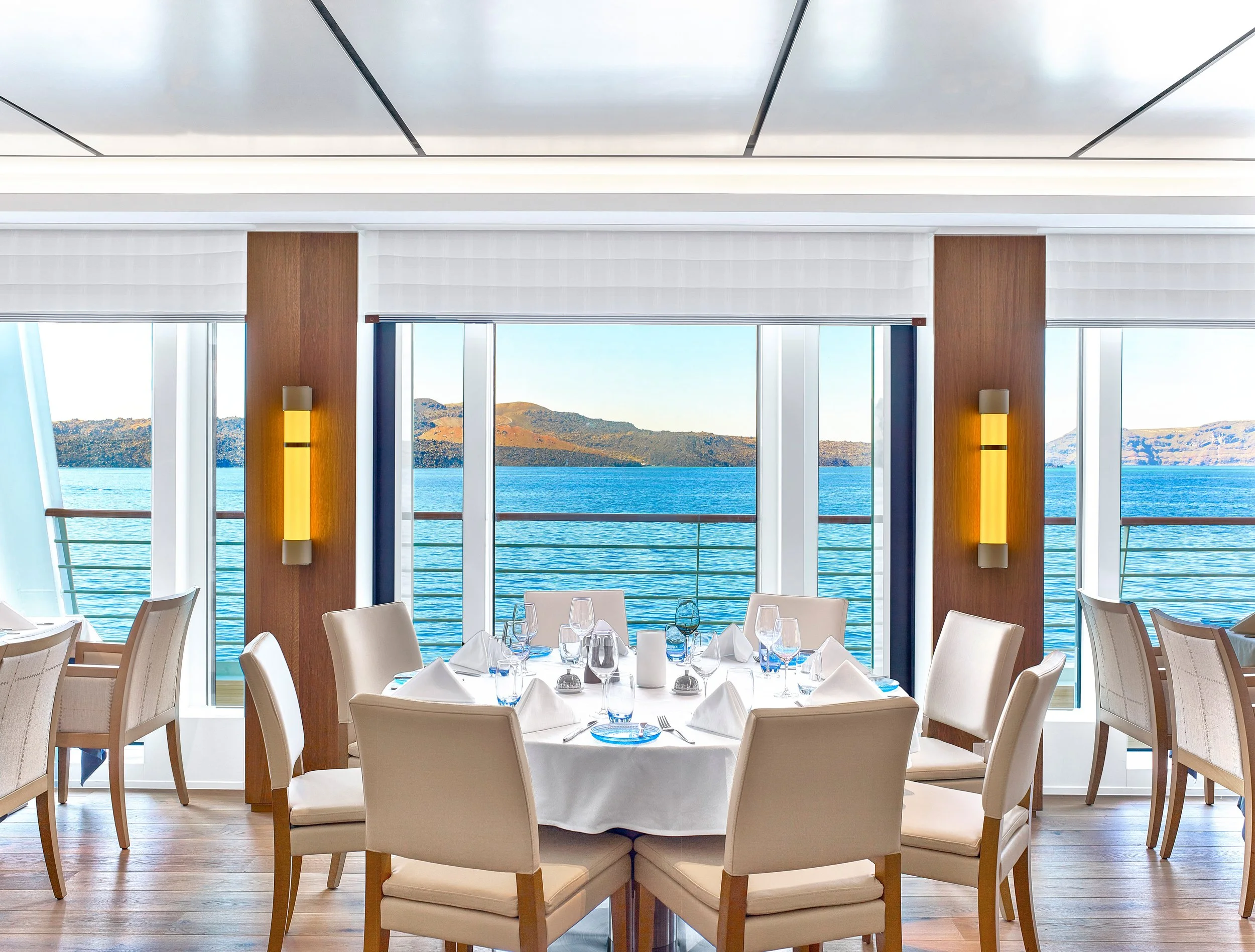 Elegant dining room with a round table set for a meal, overlooking a body of water with mountains in the background through large windows.