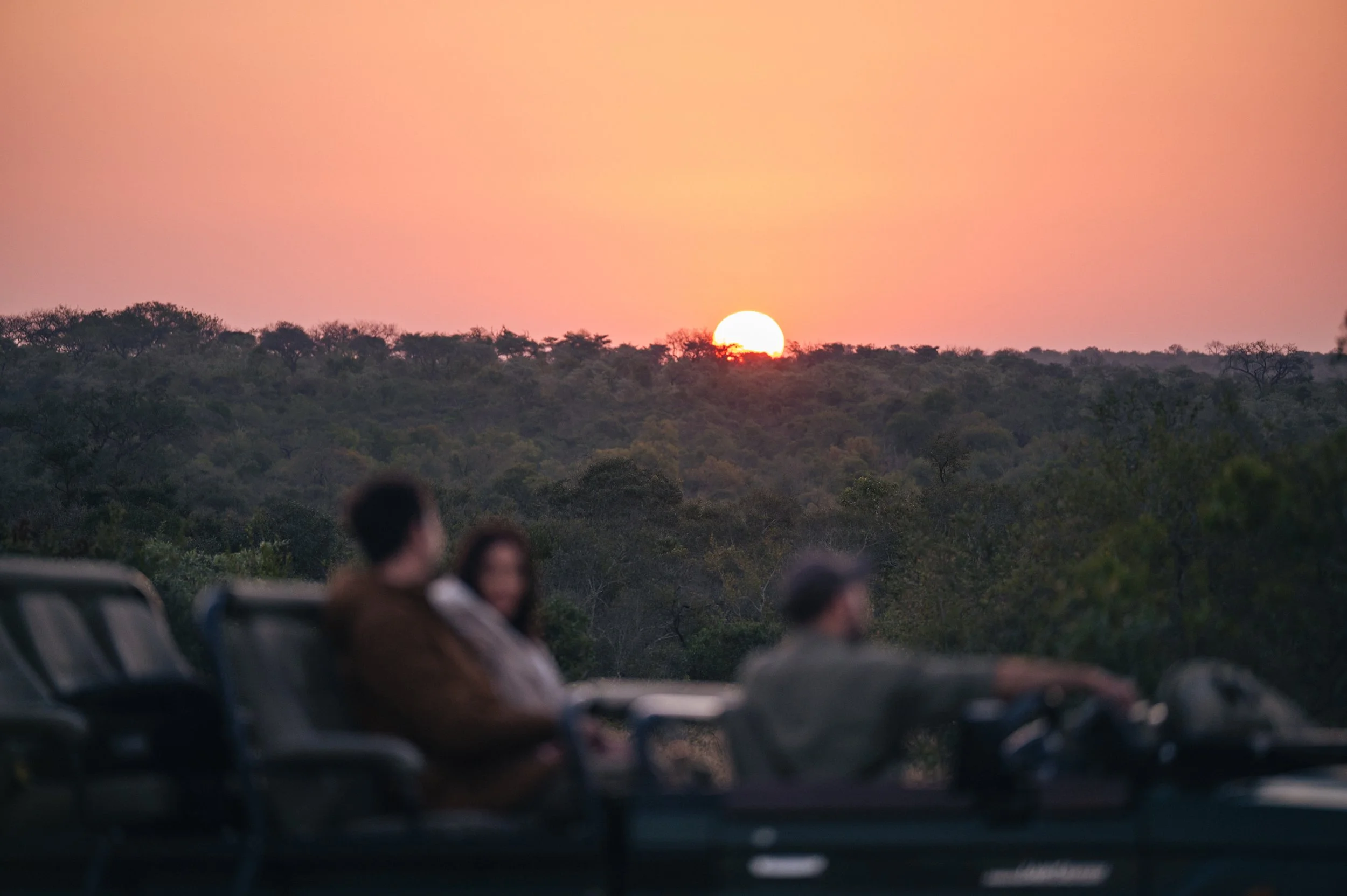 A sunset over a forested landscape with three people sitting on an open vehicle in the foreground, watching the view.