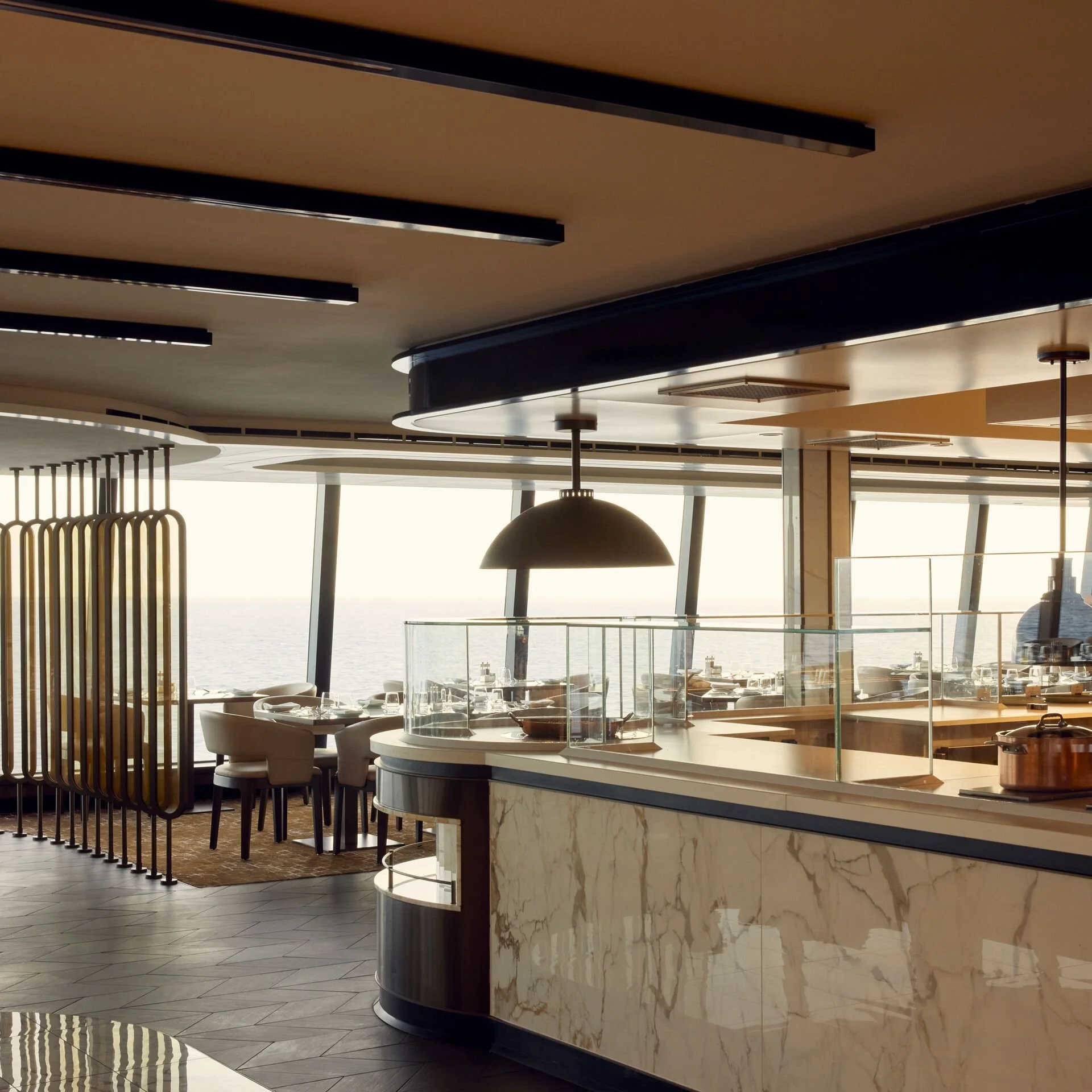 Interior of a modern restaurant with a marble counter, glass partition, and dining area with chairs and tables, overlooking the ocean through large windows.