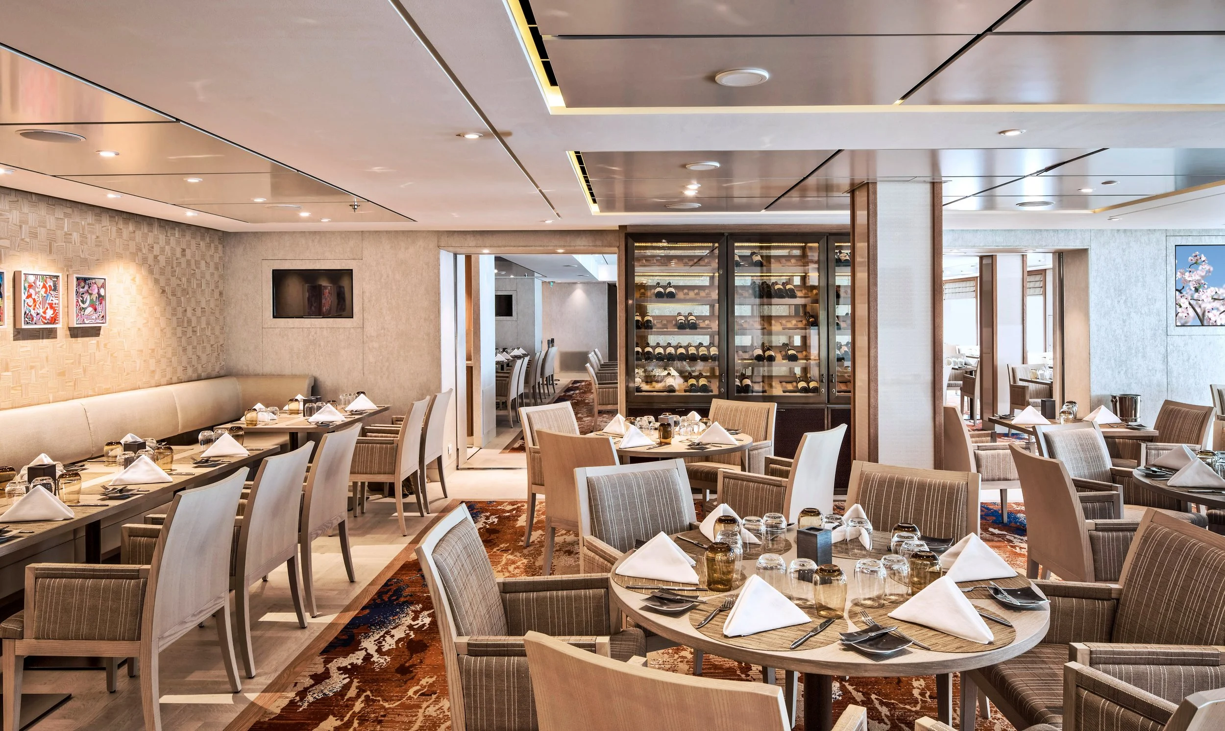 Elegant restaurant dining area with round and rectangular tables set with white napkins, glassware, and utensils. Neutral-colored chairs and a wine display case are visible, along with wall art and a television.