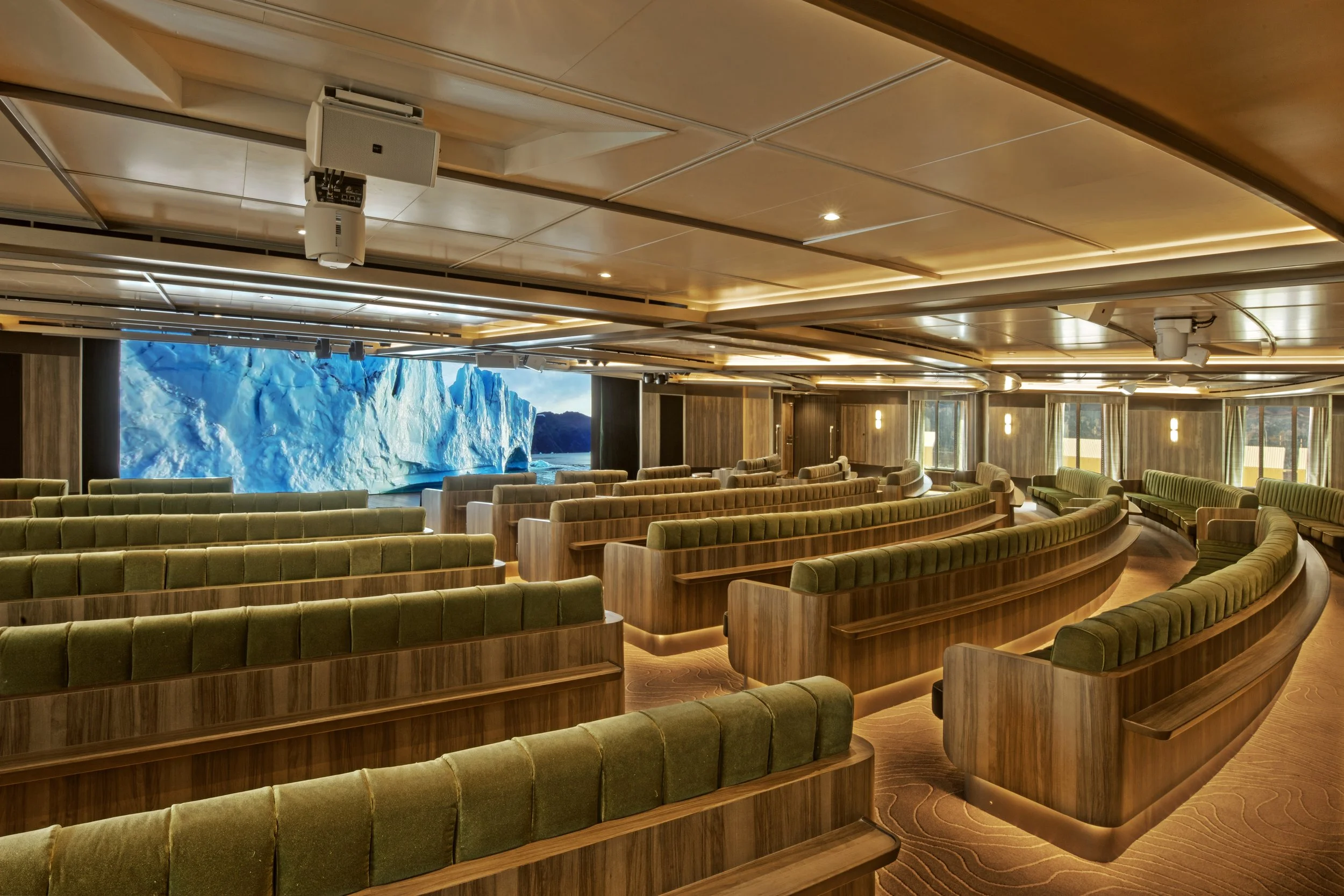 An auditorium with curved green cushioned seats arranged in tiers, a large screen displaying an iceberg, and wooden paneling with hanging lights.