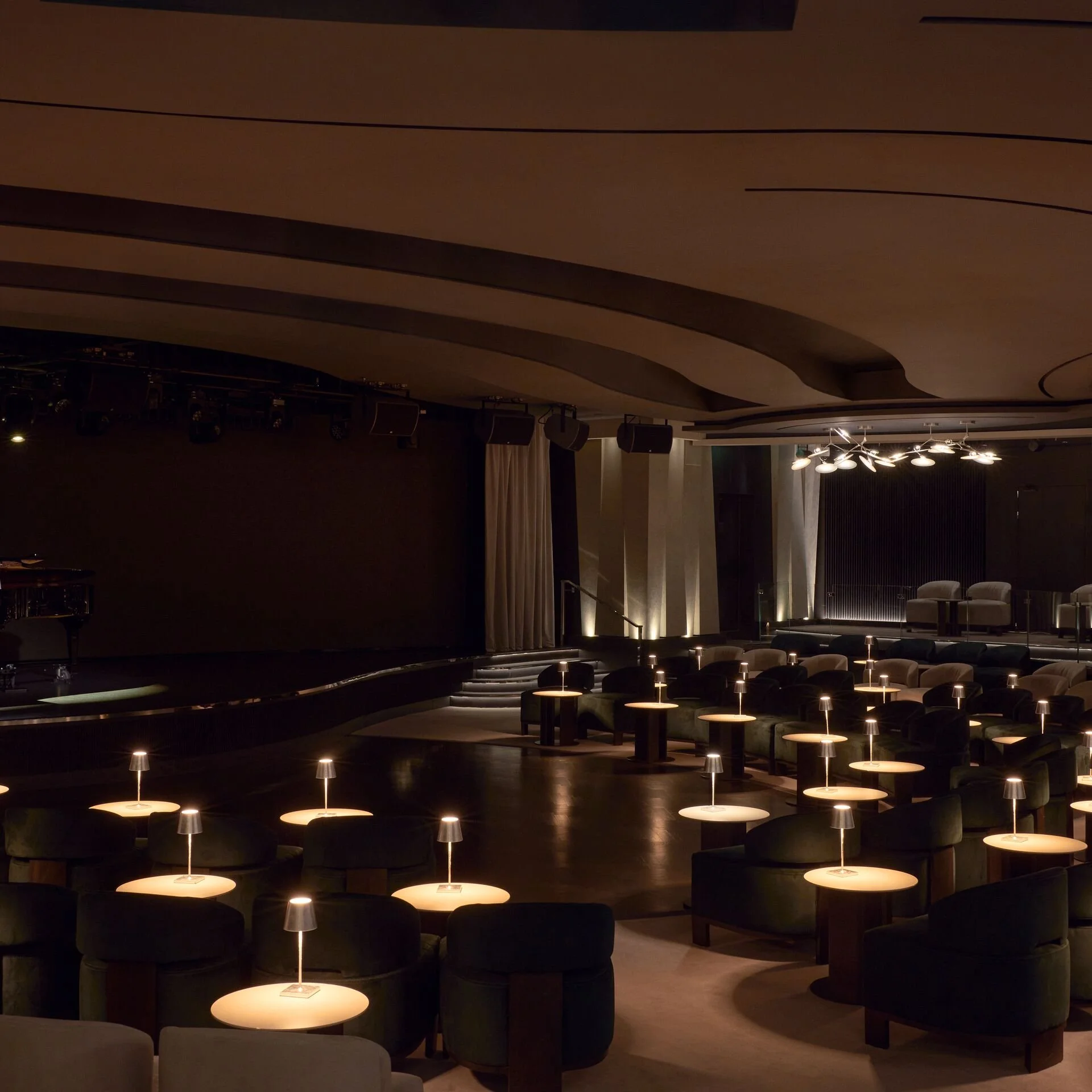 Empty theater or lounge with small tables and lamps, stage and seating area in background, dim lighting.