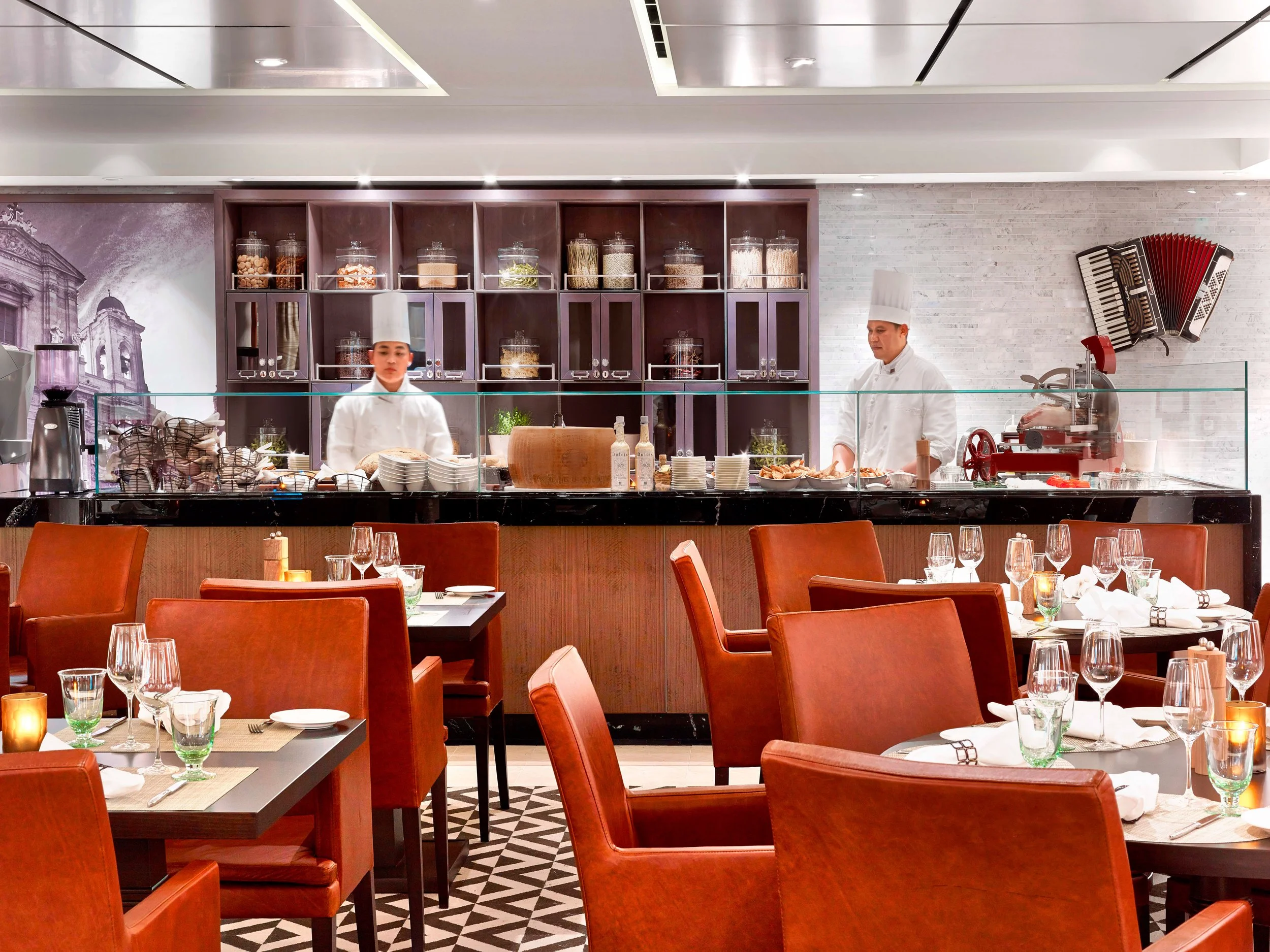 Elegant restaurant interior with tables set with glassware and napkins, a buffet area with chefs preparing food in the background, and kitchen tools on the wall.