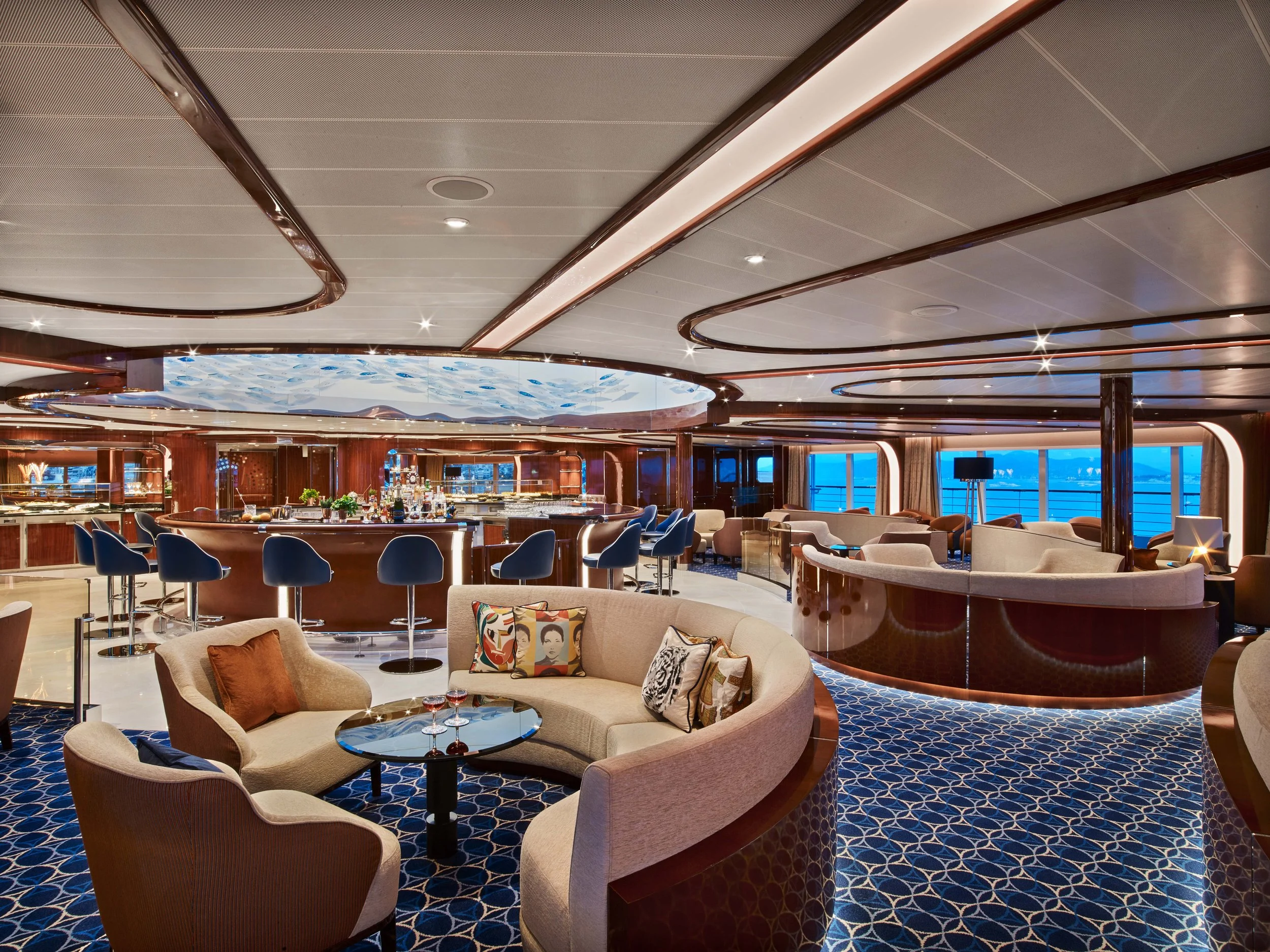Elegant lounge area on a cruise ship with a bar, barstools, seating, and large windows overlooking the ocean.