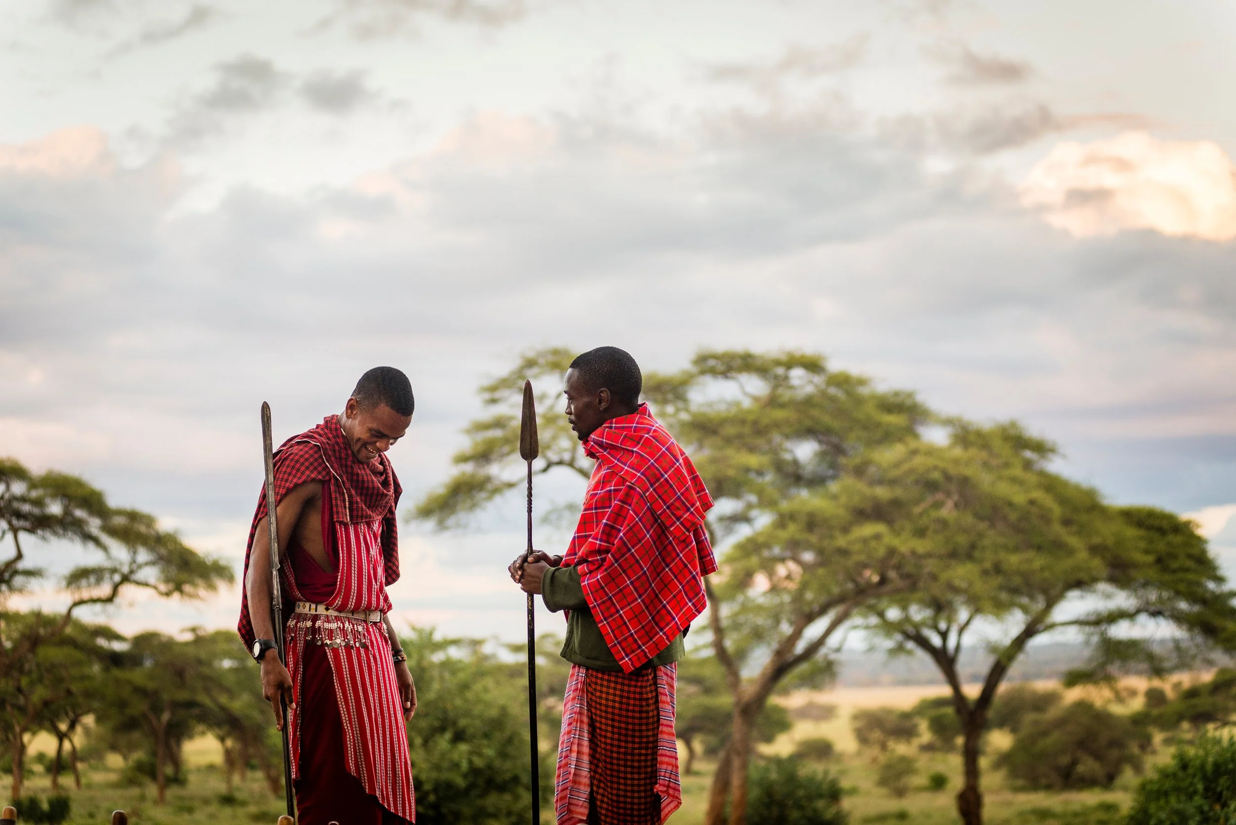 Two Maasai men in traditional red and black shuka clothing standing outdoors among trees, with one holding a spear and the other looking down and smiling, under a cloudy sky.