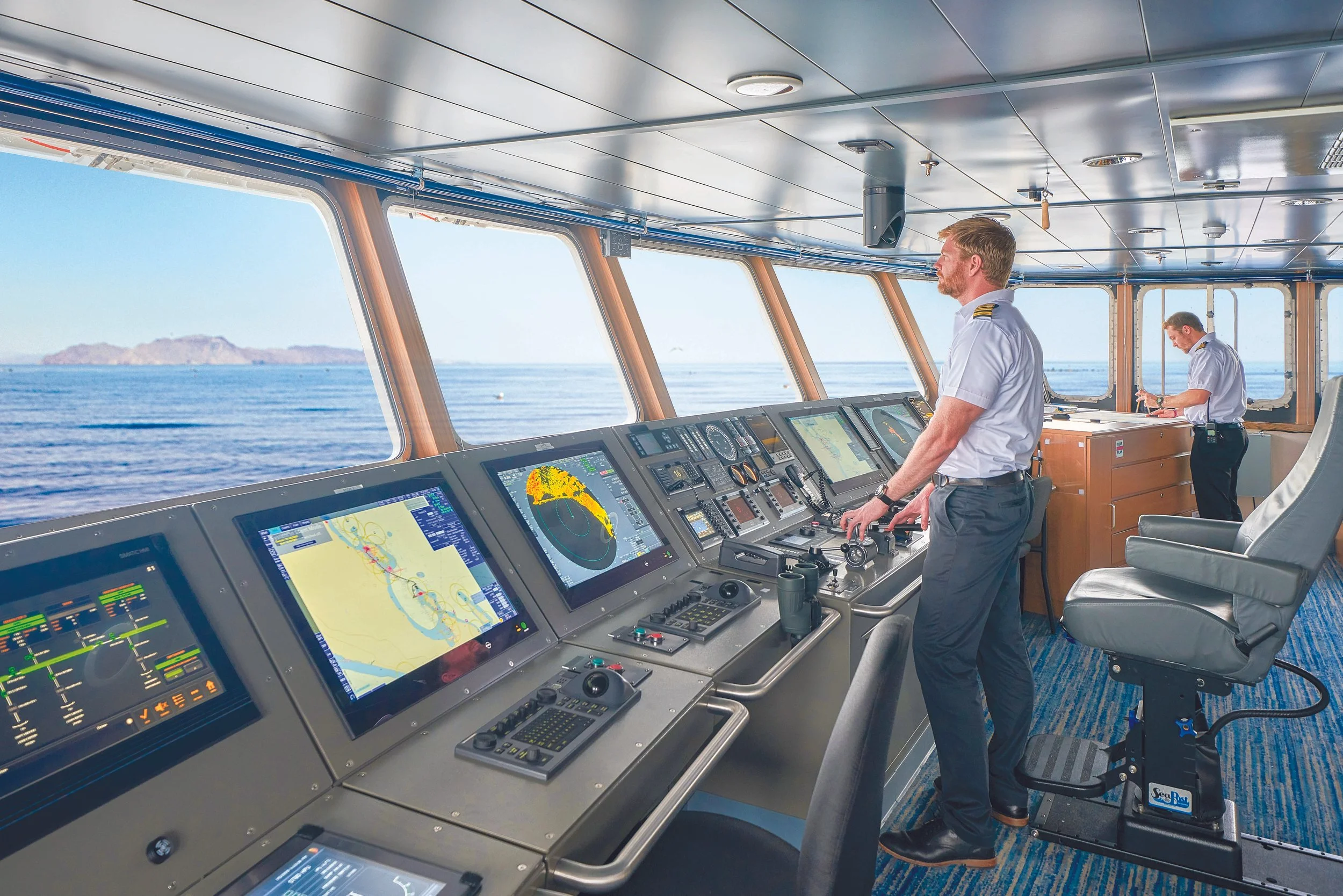 Two ship officers in uniform working at the ship's helm in a control room with large windows showing the ocean and islands outside.