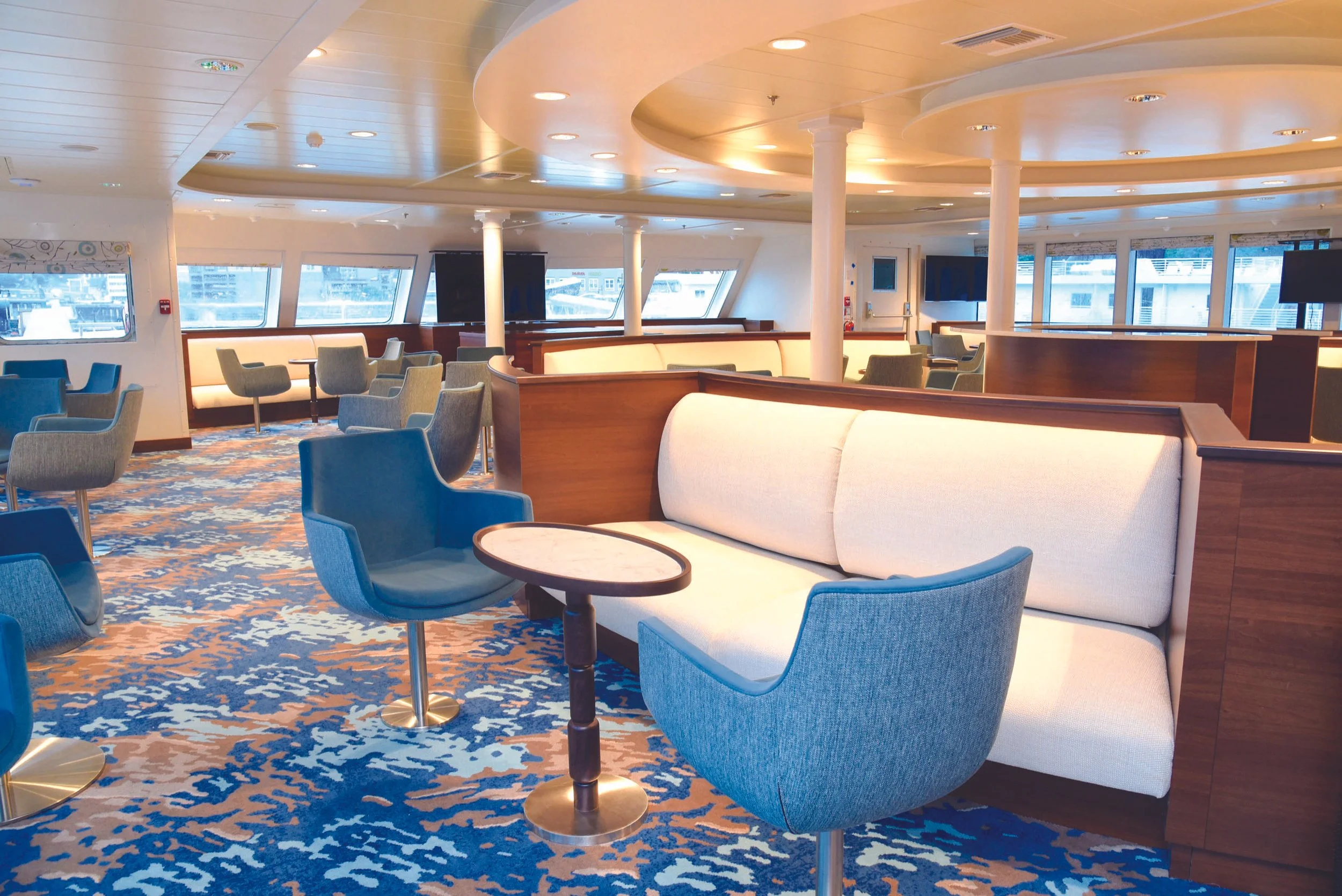 Empty lounge area on a cruise ship with blue and beige chairs, white and beige sofas, small wooden tables, large windows with a view of the harbor, and a colorful patterned carpet.