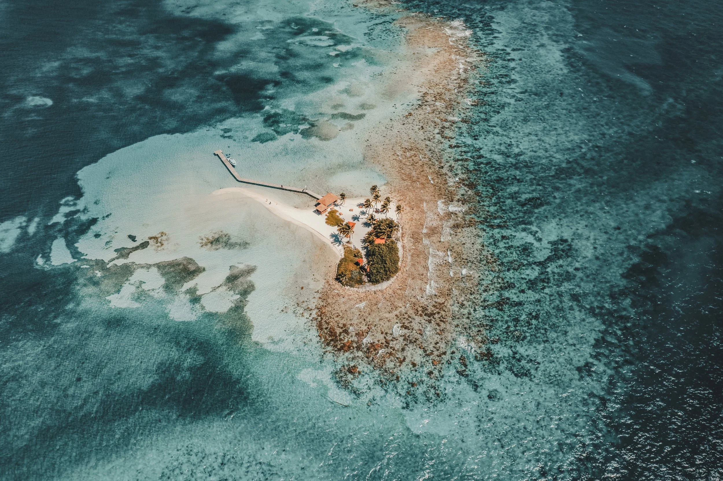 Aerial view of a small tropical island with a dock, palm trees, and a white sandy beach surrounded by turquoise ocean water.
