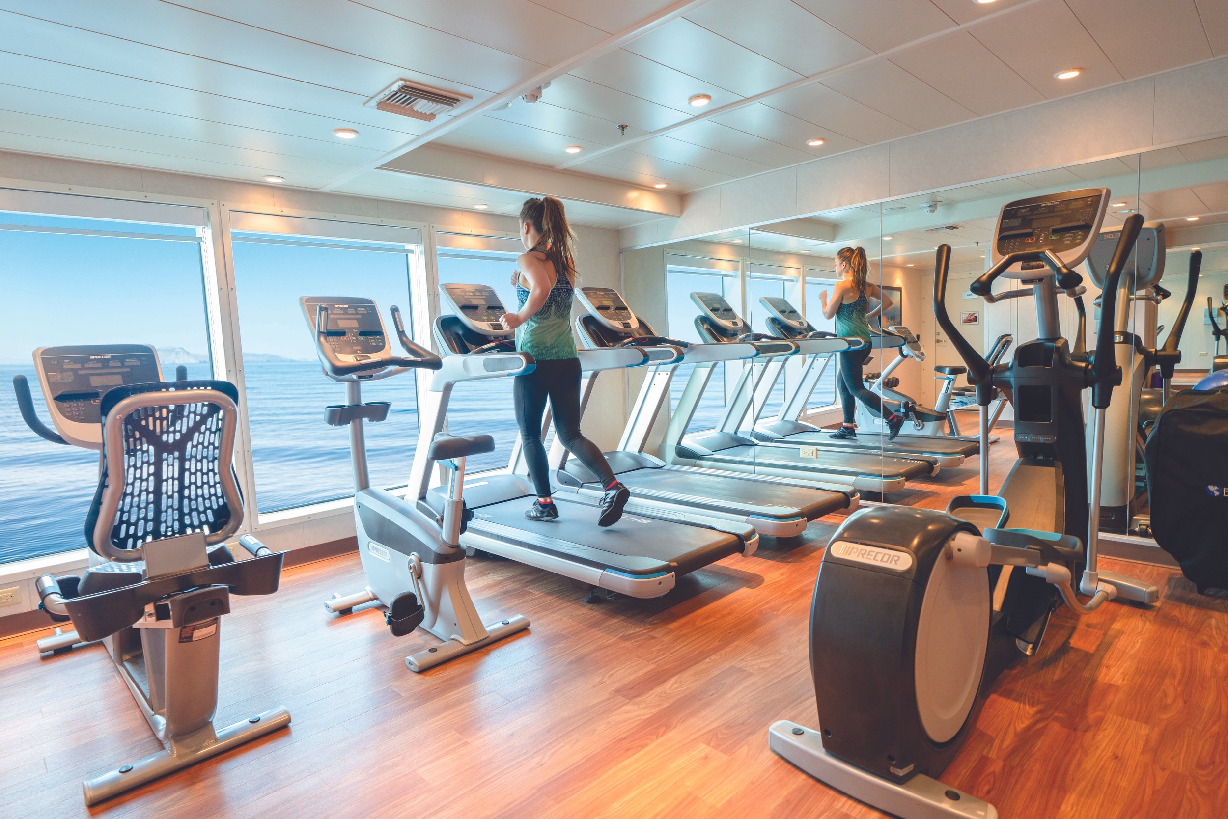Interior of a gym on a cruise ship with cardio equipment, including treadmills, an elliptical, and stationary bikes, facing large windows overlooking the ocean. Two women are running on the treadmills.