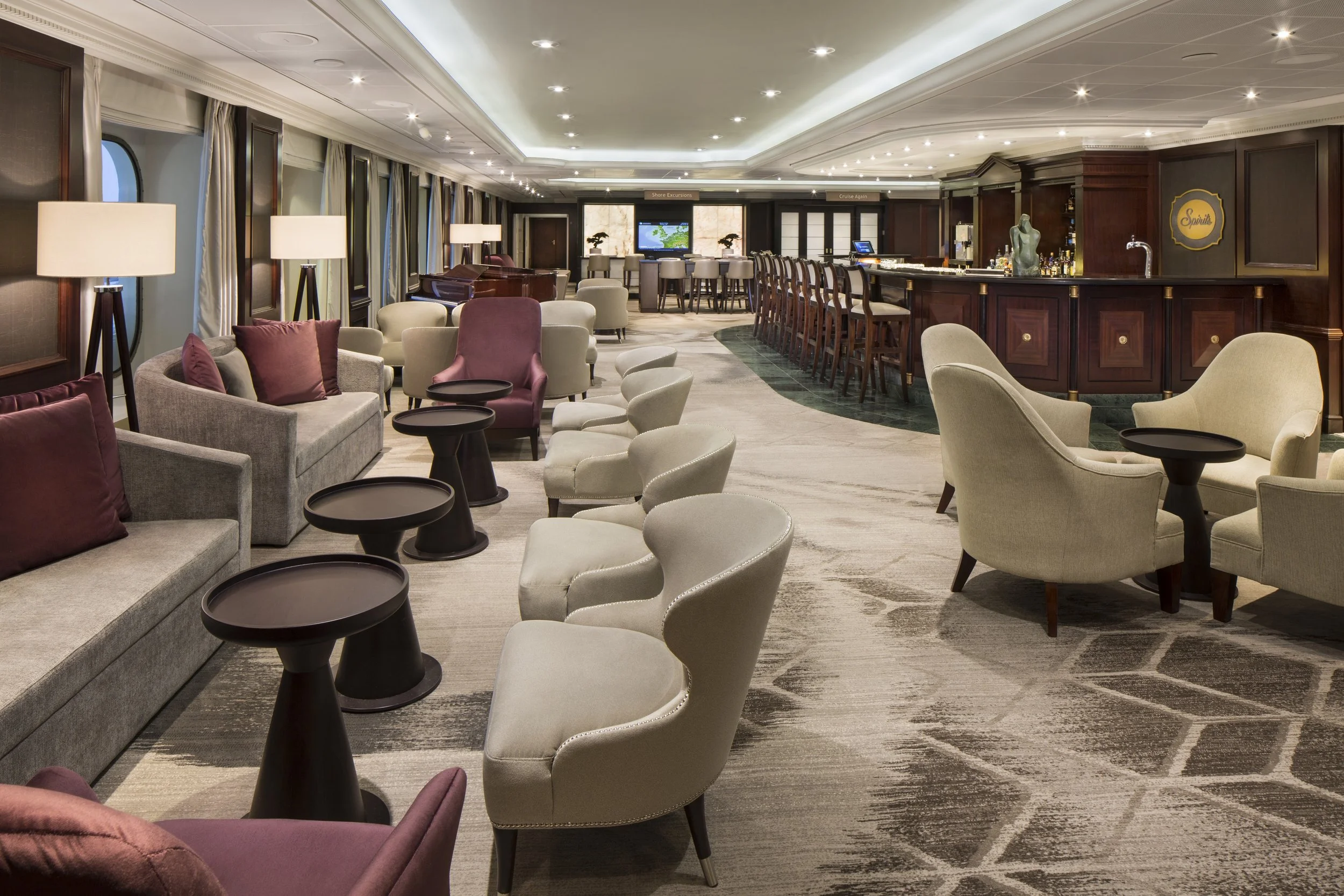 A luxurious lounge area with plush chairs and sofas, small black tables, a bar with high stools, and a large flat screen TV in the background, illuminated by overhead recessed lighting.