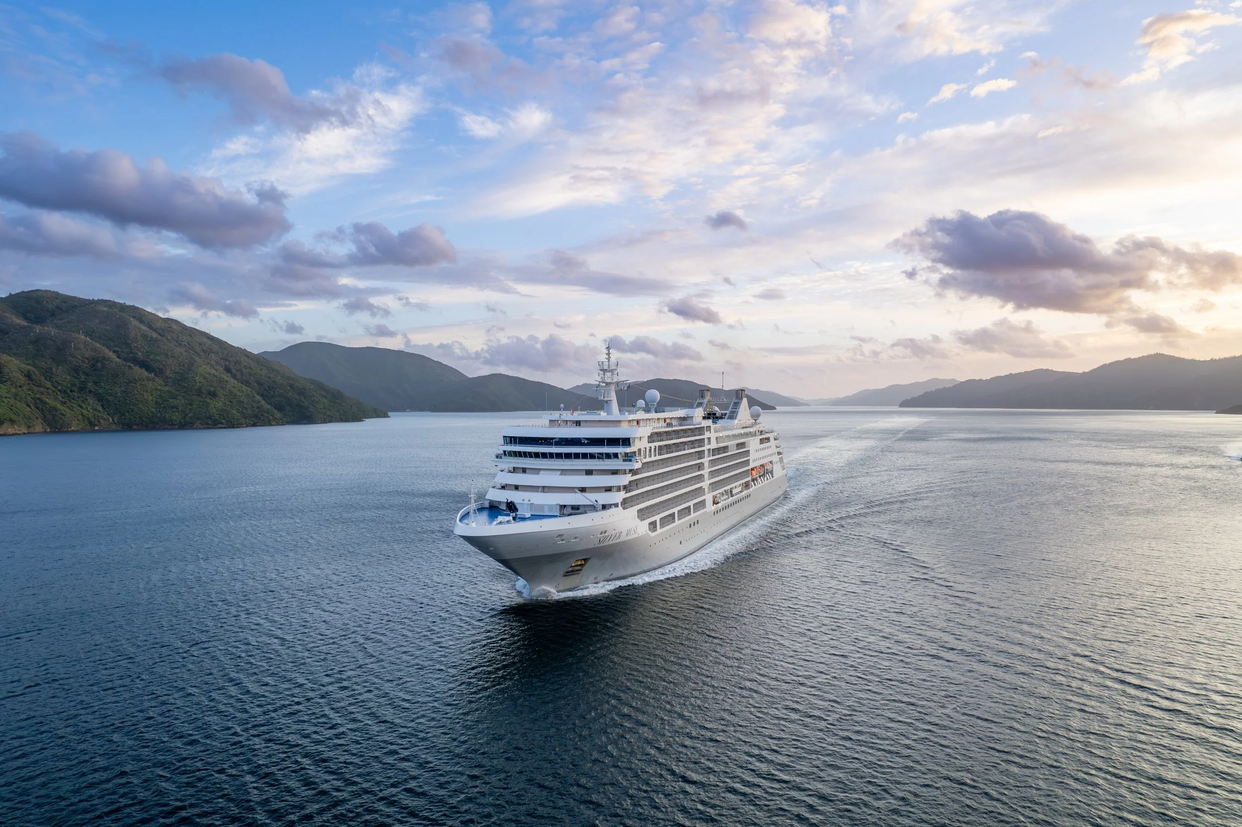 A large white cruise ship sailing on a calm body of water with green hills on both sides and a partly cloudy sky with the sun setting or rising.
