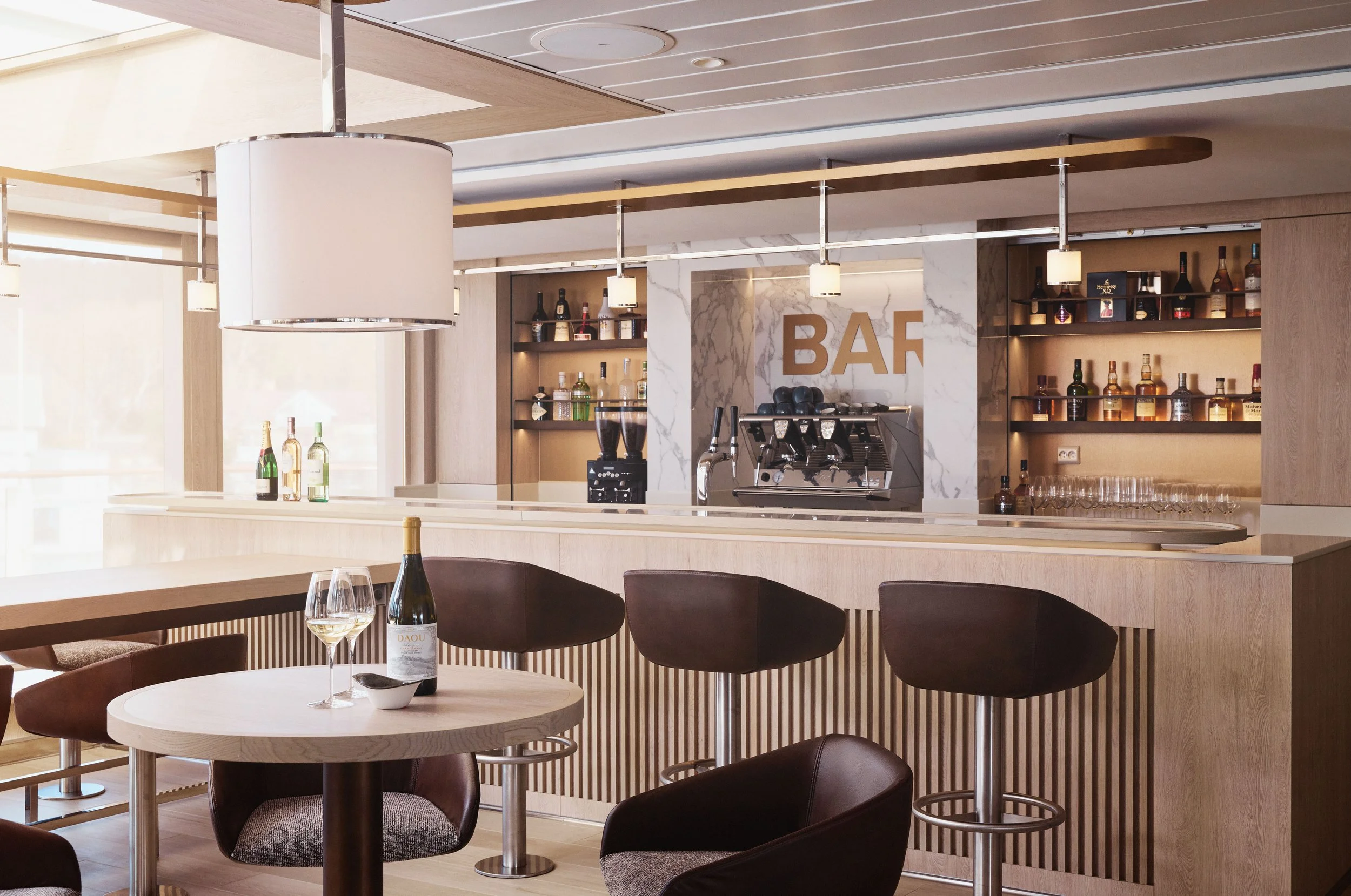 Modern bar with white wood and marble accents, bottles of alcohol on shelves, coffee machine, and a table with a bottle of white wine and two glasses in a well-lit space.