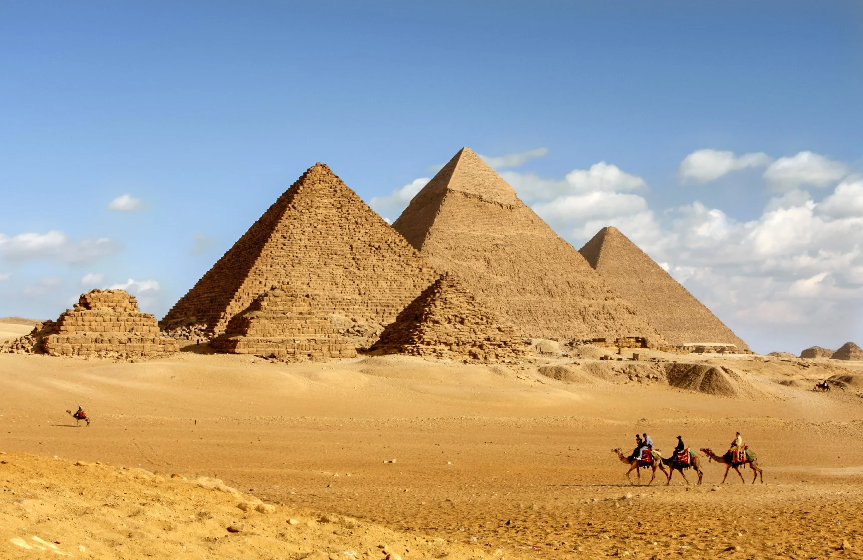 The image shows the pyramids of Egypt in a desert landscape with a few people riding camels in the foreground.