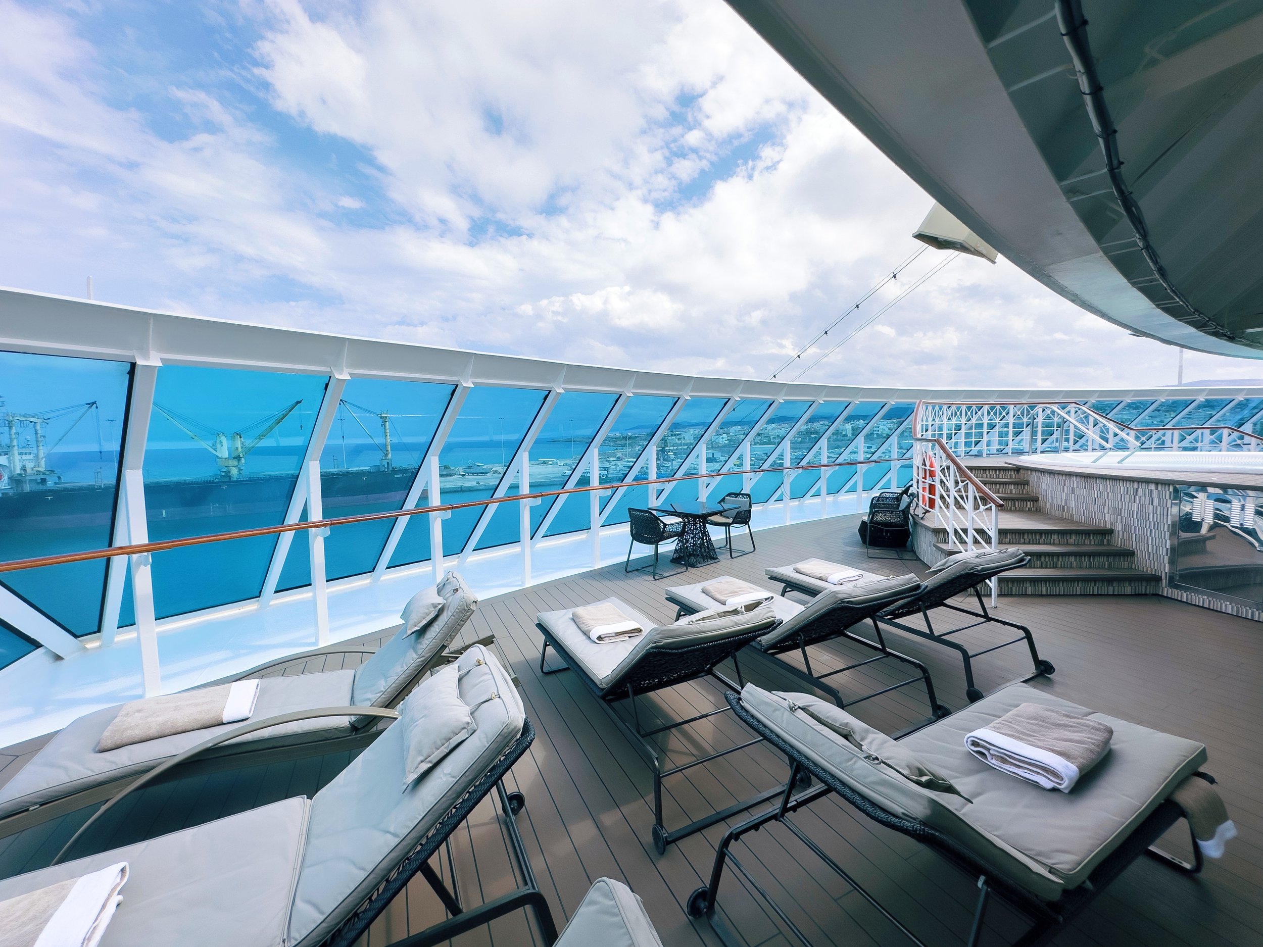 View of a cruise ship deck with lounge chairs, towels, and a glass barrier, overlooking a harbor under a partly cloudy sky.