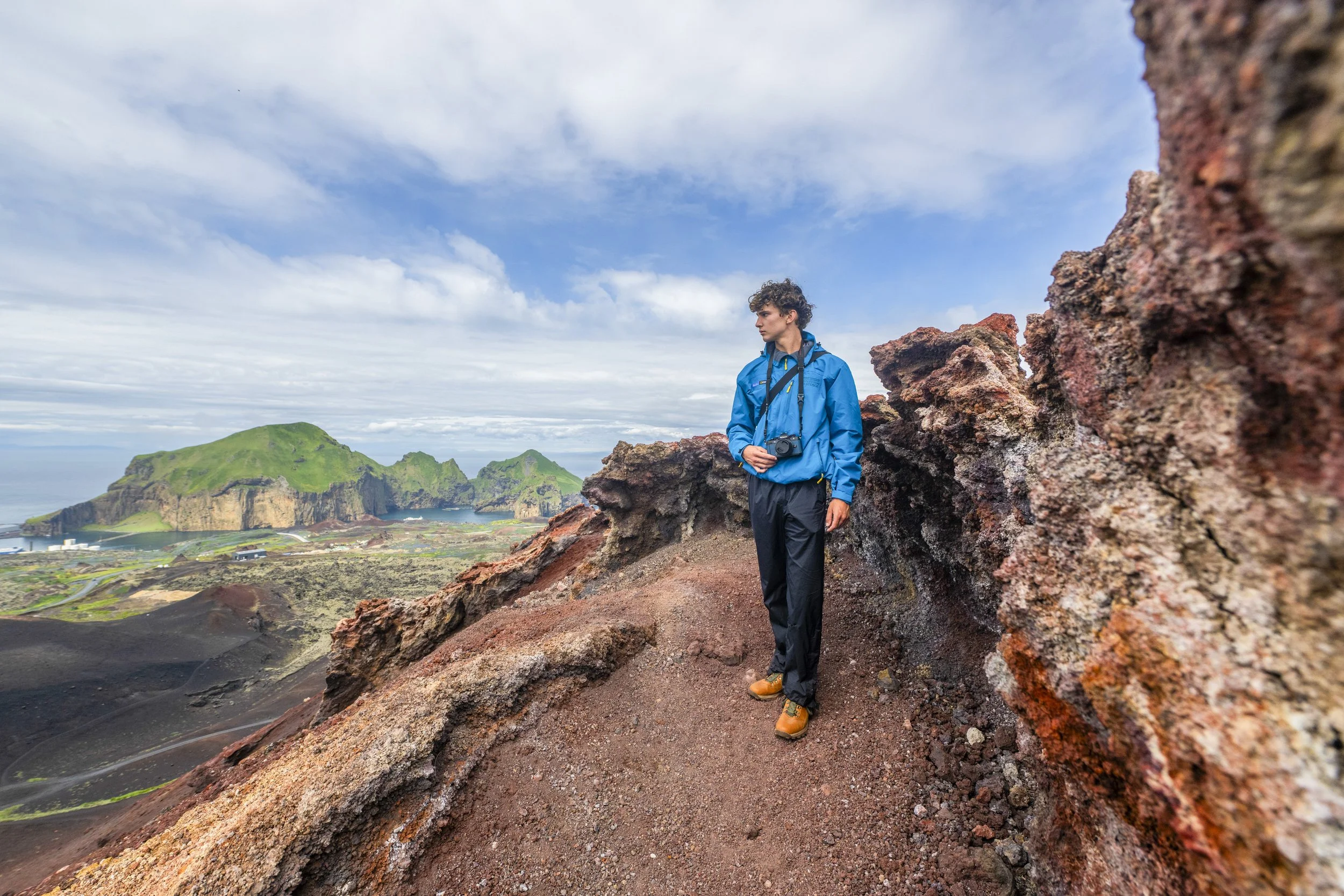 Young man in blue jacket, black pants, and hiking boots standing on volcanic rock with a camera around his neck, overlooking a landscape with green hills and ocean under a cloudy sky.