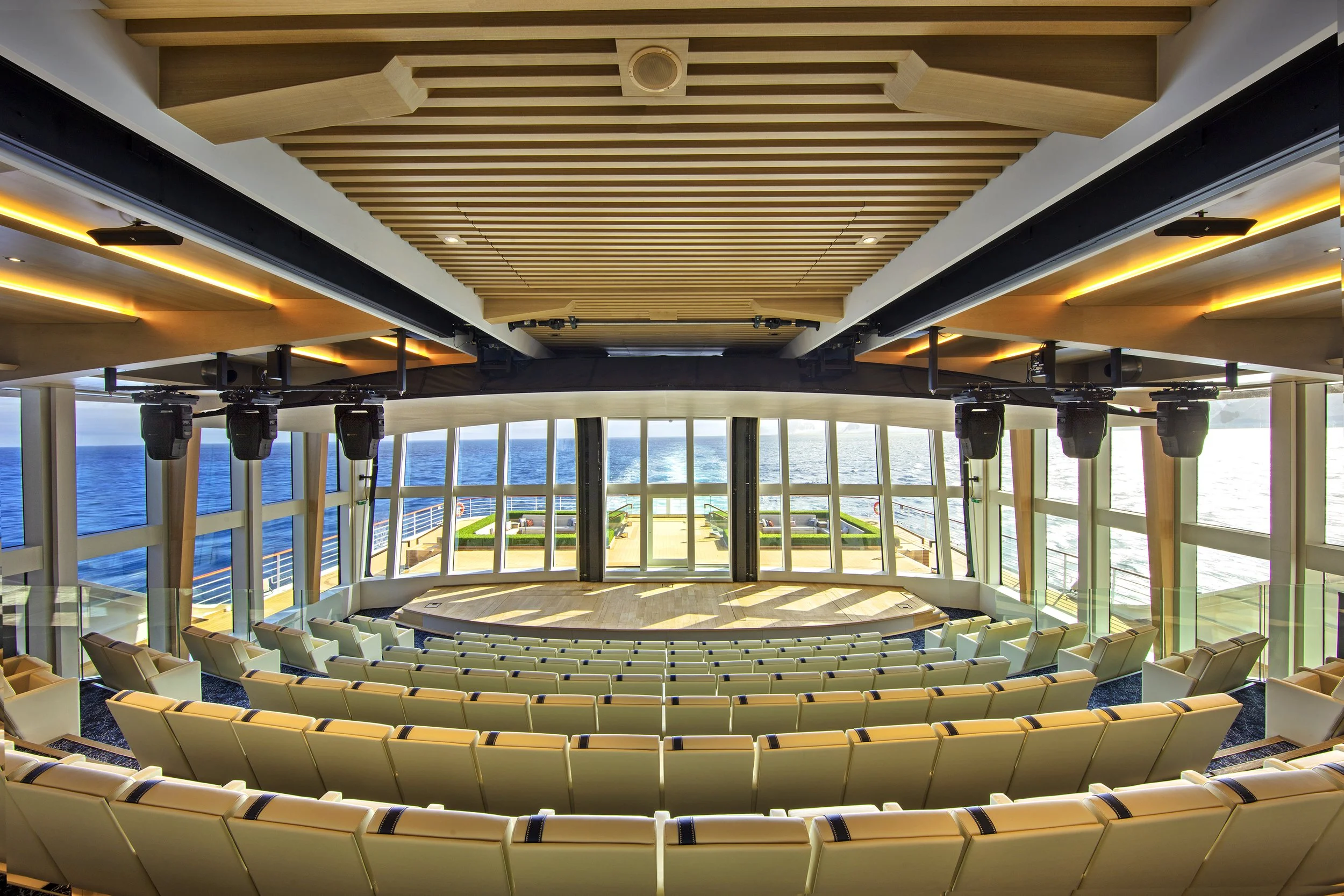 The interior of a modern auditorium or theater on a cruise ship with rows of cream-colored seats facing a stage, large windows showing the ocean, and ceiling with wood paneling and stage lighting equipment.