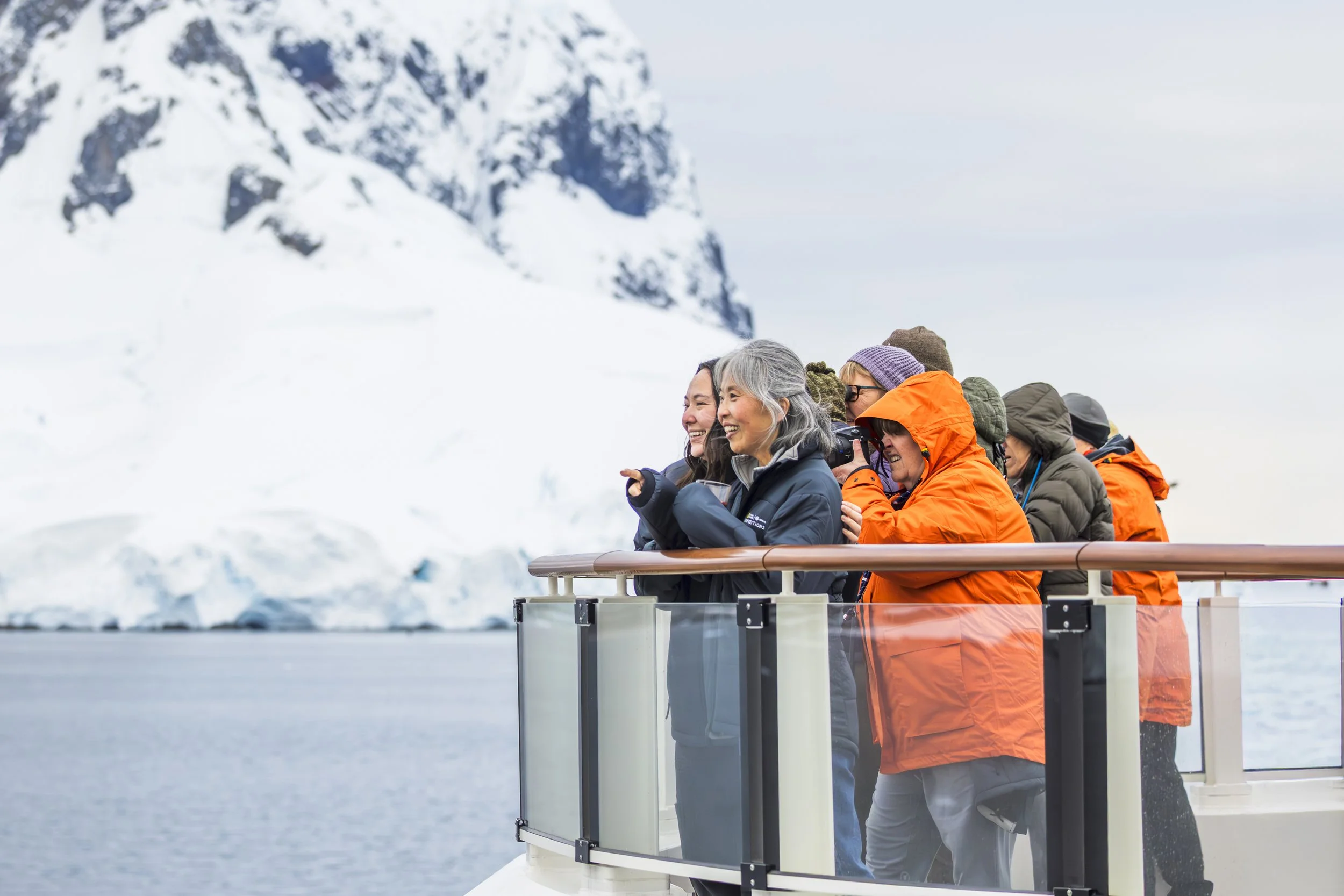 Group of people on a boat deck looking at snowy mountains and icebergs, dressed in winter clothing, including orange and gray jackets.