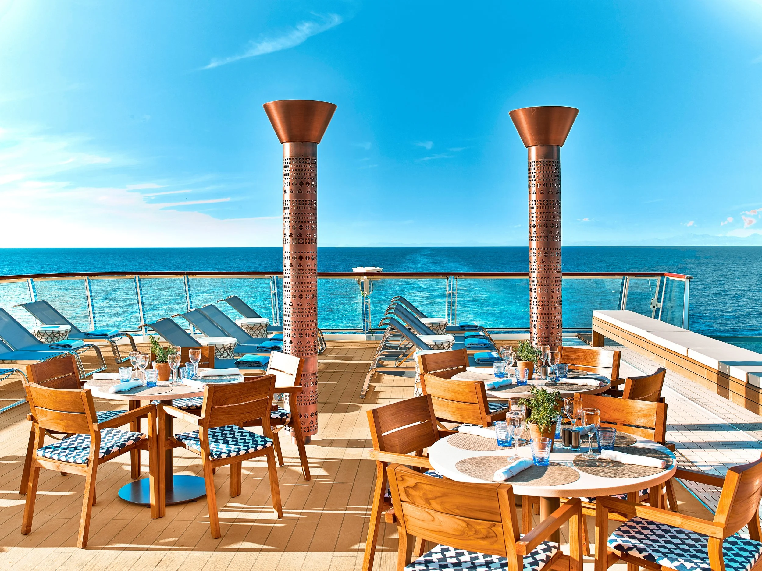 Outdoor dining and lounging area on a cruise ship deck overlooking the ocean, with tables set for a meal, sun loungers, and two large decorative columns.