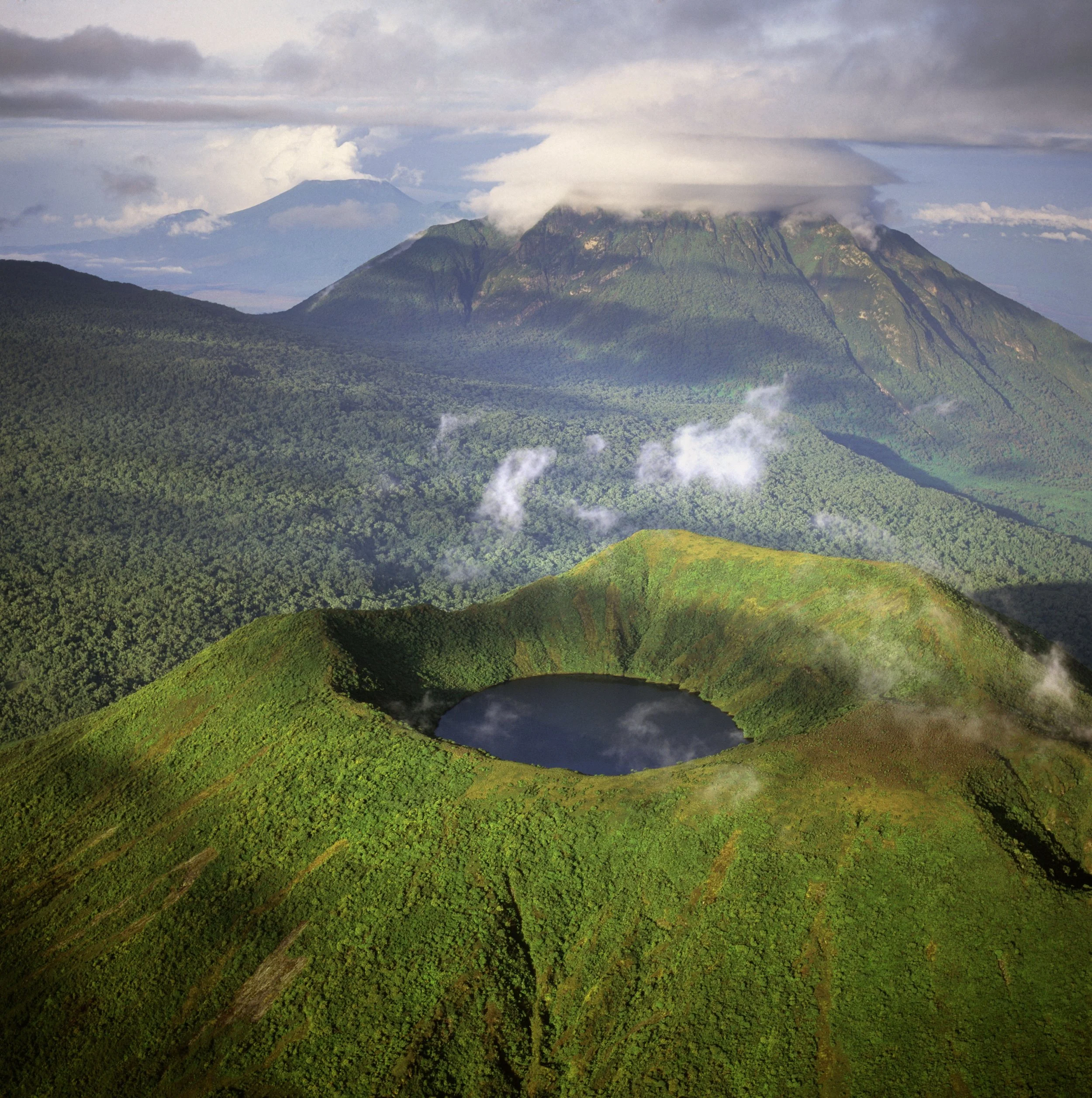 A volcanic landscape with a crater lake in the foreground, surrounded by green hills, and a mountain with a cloud-covered peak in the background.