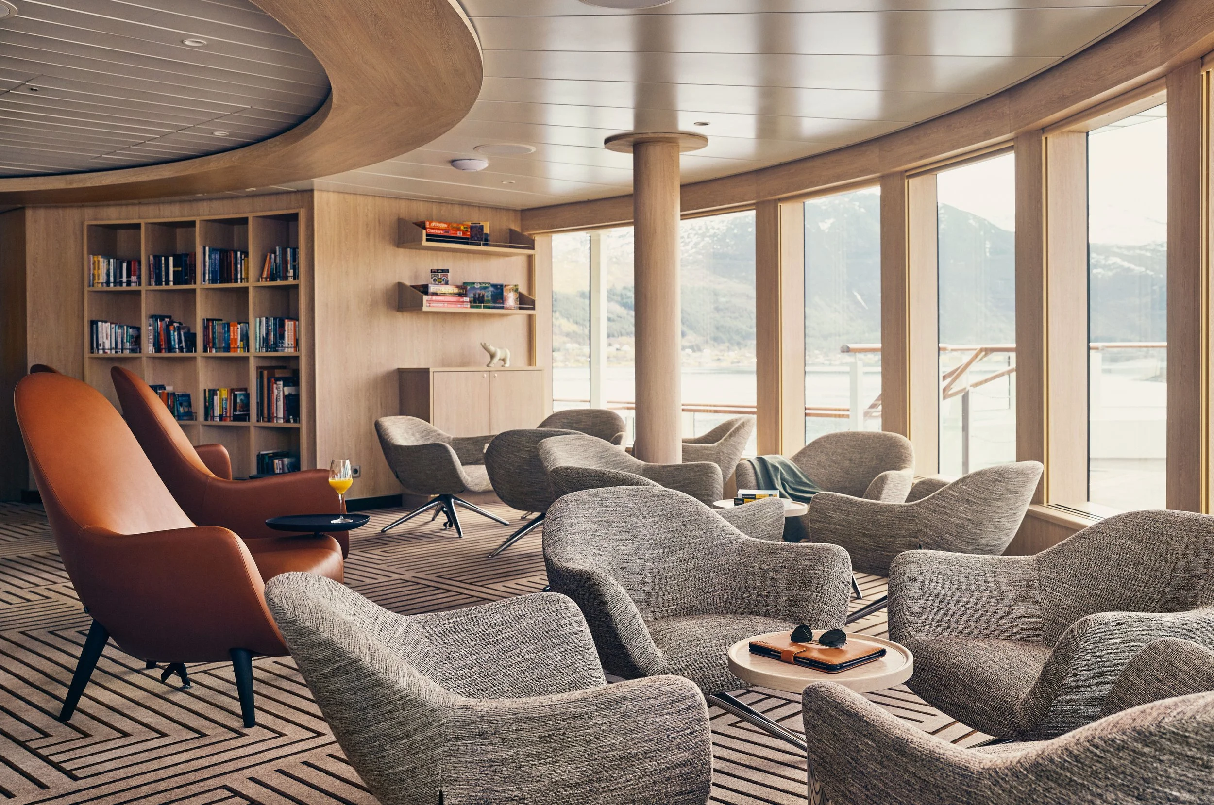 A modern lounge with multiple gray armchairs, two orange high-back chairs, a glass of orange drink on a small black side table, a bookshelf filled with books on the left, and large windows with a view of mountains and water outside.