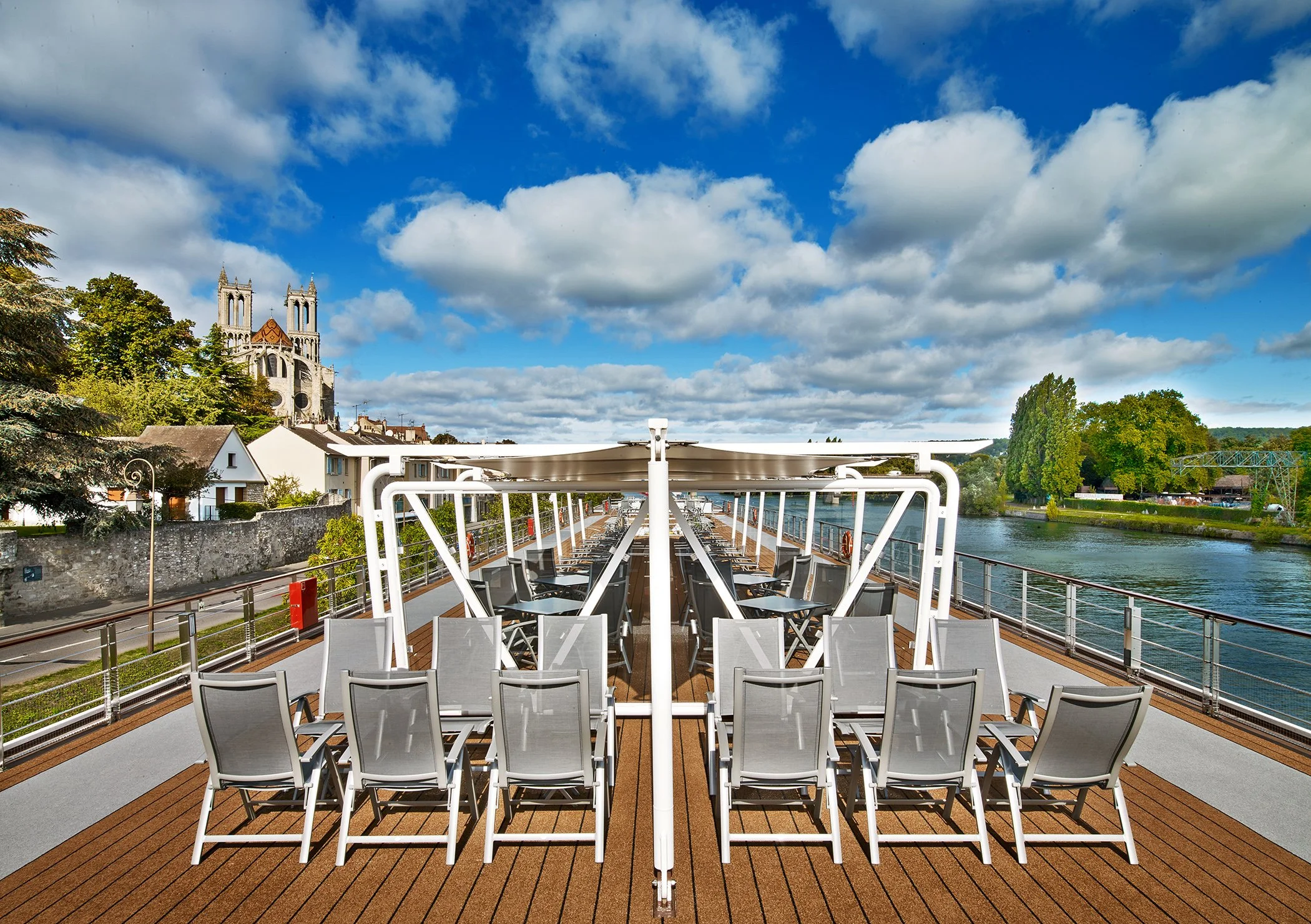 Empty outdoor deck with rows of gray chairs, white railings, and a river on the side, under a sky with clouds and distant trees and buildings, including a church with twin spires.