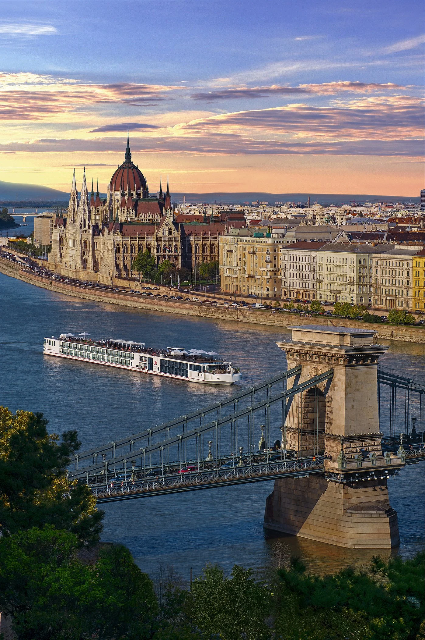 View of the Hungarian Parliament building along the Danube River in Budapest, Hungary during sunset, with the Chain Bridge in the foreground.