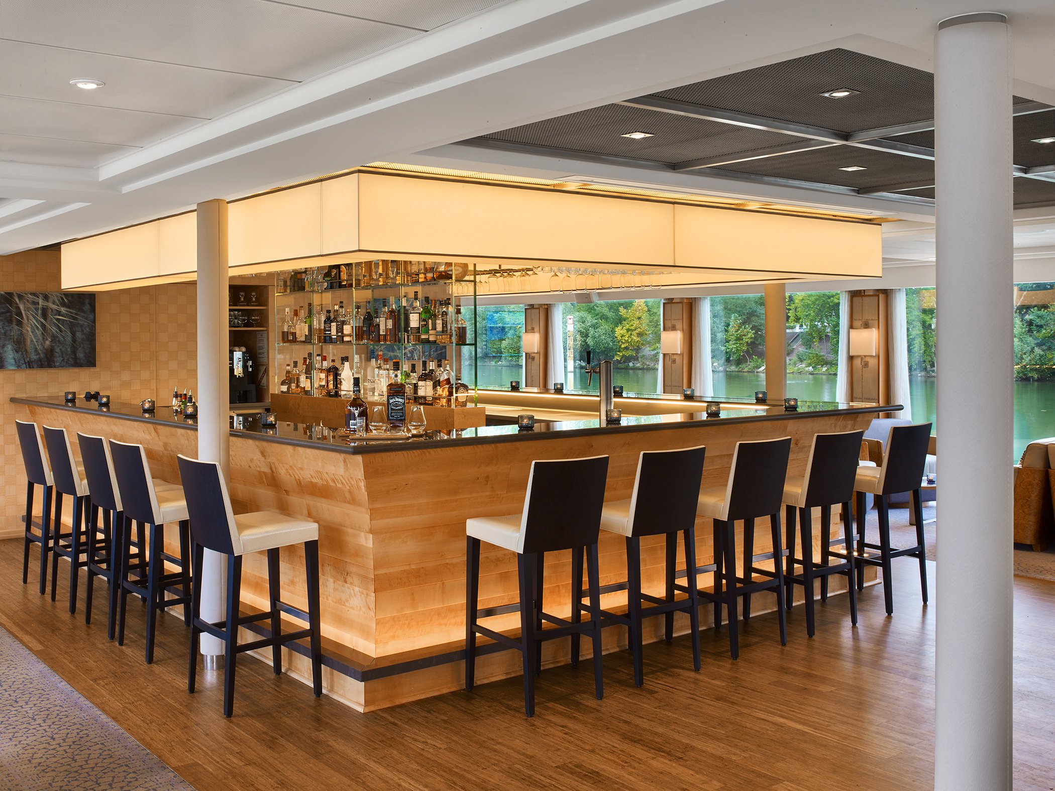 Modern bar with a wooden counter, black and white chairs, bottles of alcohol on shelves, large windows with a river view outside.