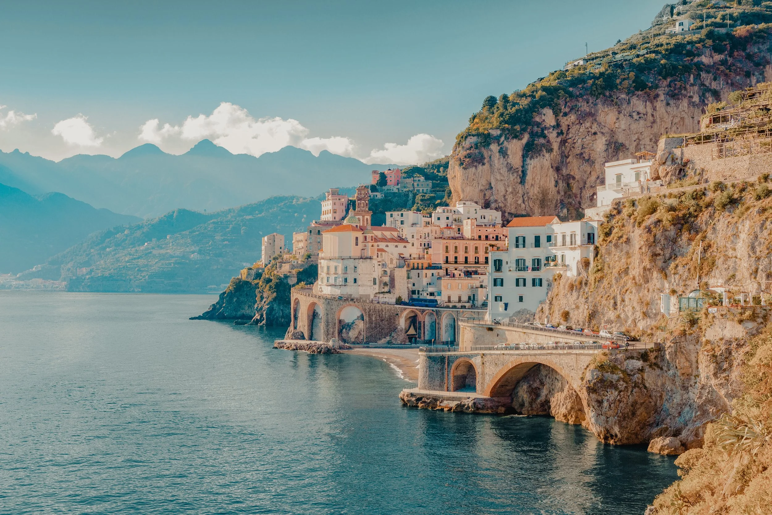 Coastal town built into cliffs along a calm sea, with mountains in the background, featuring colorful buildings and an arched stone bridge over the water.