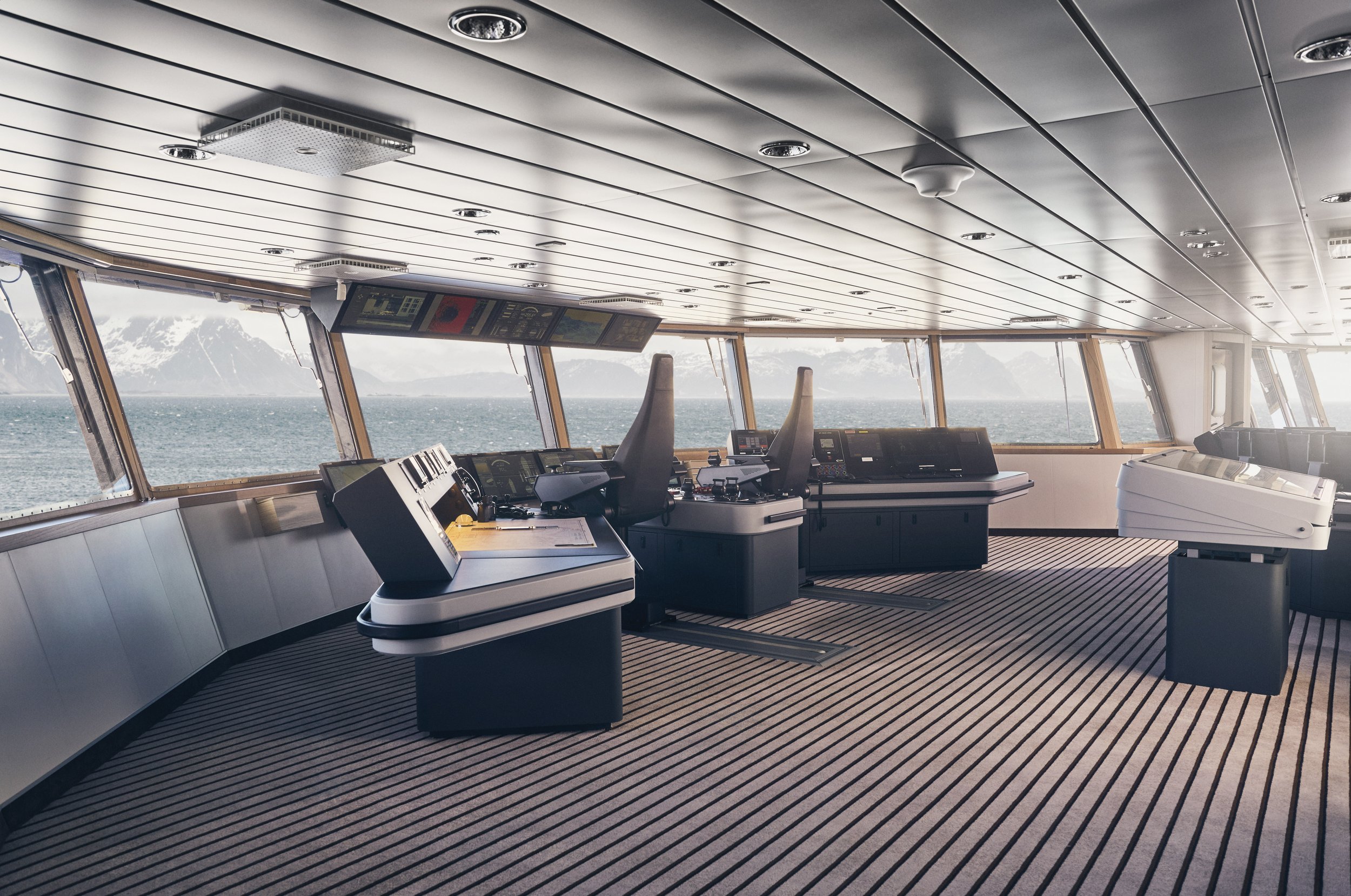 Bridge of a ship with navigation and control equipment, large windows showing water and mountains outside, striped carpet, and chairs at the helm station.