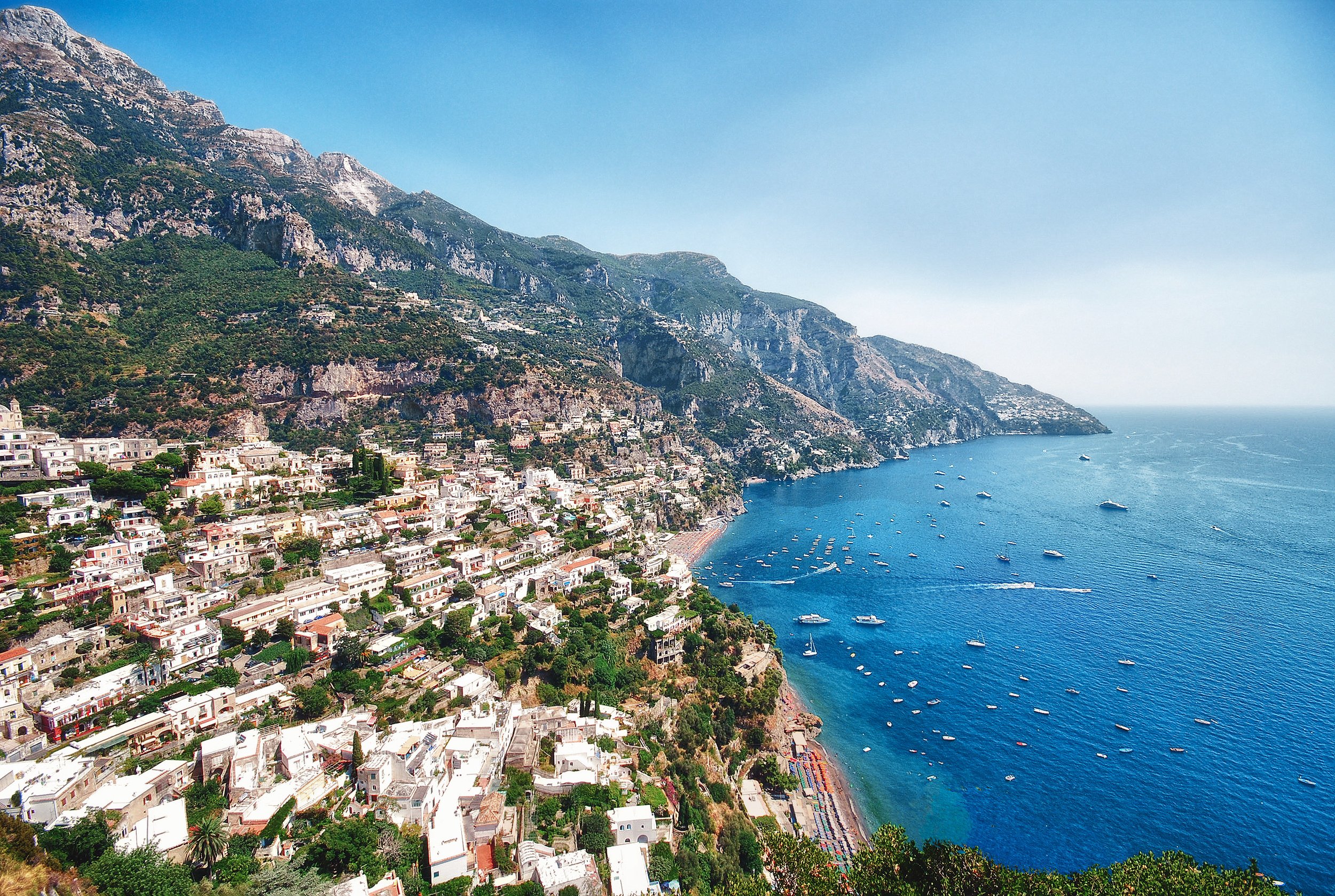 A coastal village on a hillside with white buildings and narrow streets, overlooking the blue sea with boats and yachts, and green mountains in the background.