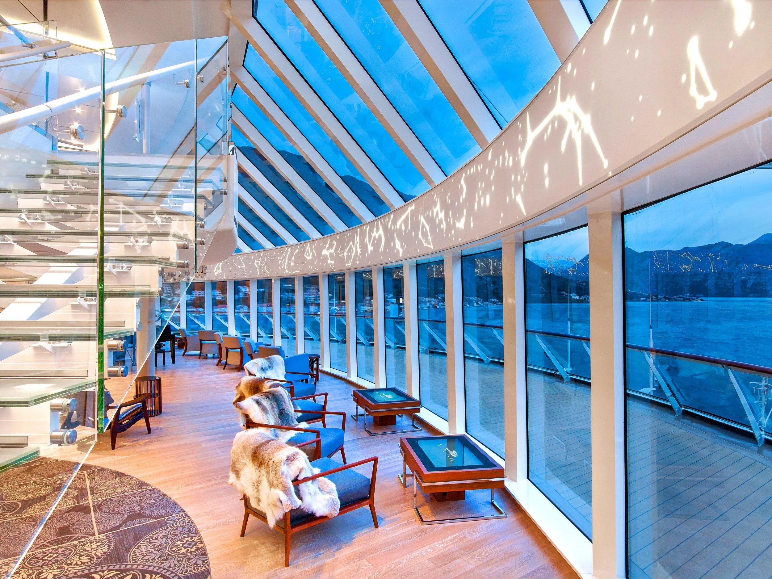 Interior of a modern building with large floor-to-ceiling glass windows showing a scenic mountain view; the space features wooden flooring, chairs with fur throws, and decorative tables.