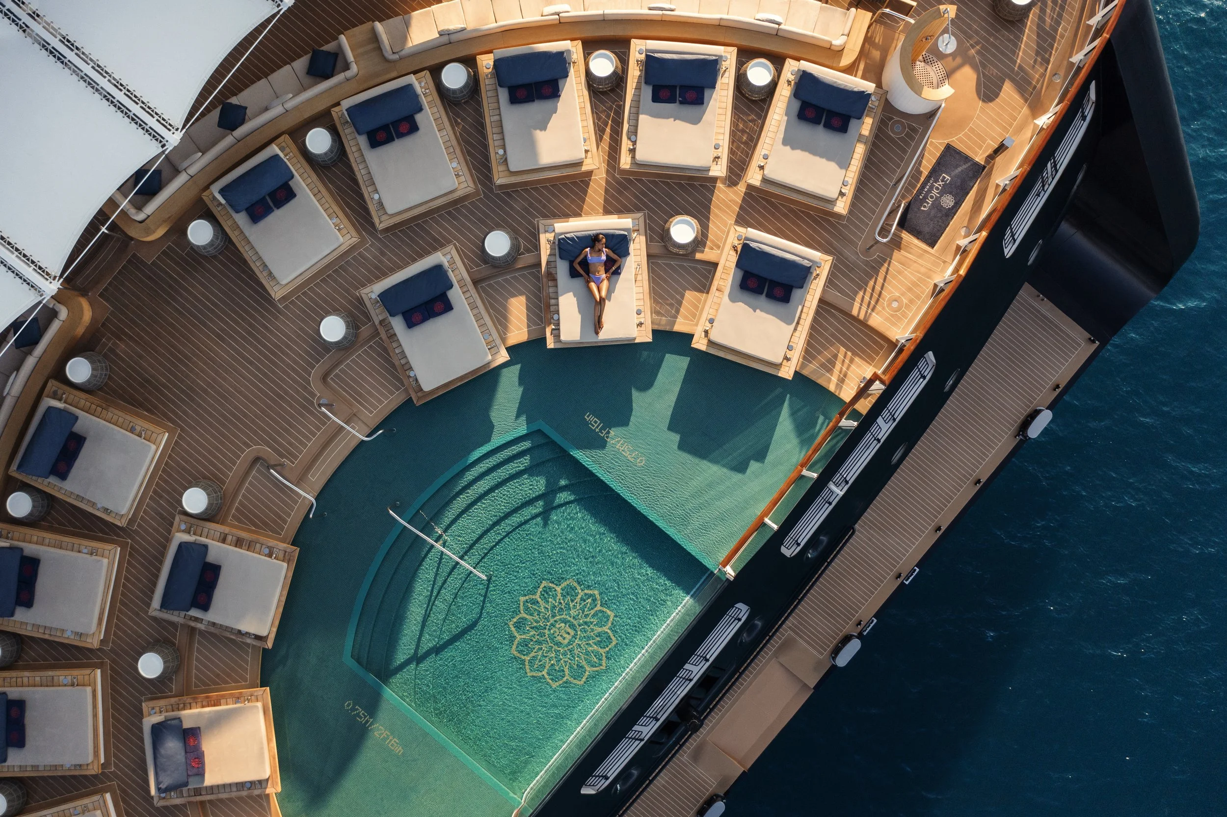 Aerial view of a cruise ship's outdoor pool area with a woman lounging on a sunbed near the pool. The pool has a decorative design at the bottom, and surrounding deck chairs with blue cushions are arranged along the wooden deck. The ship is on the water, with part of the ocean visible on the right side.
