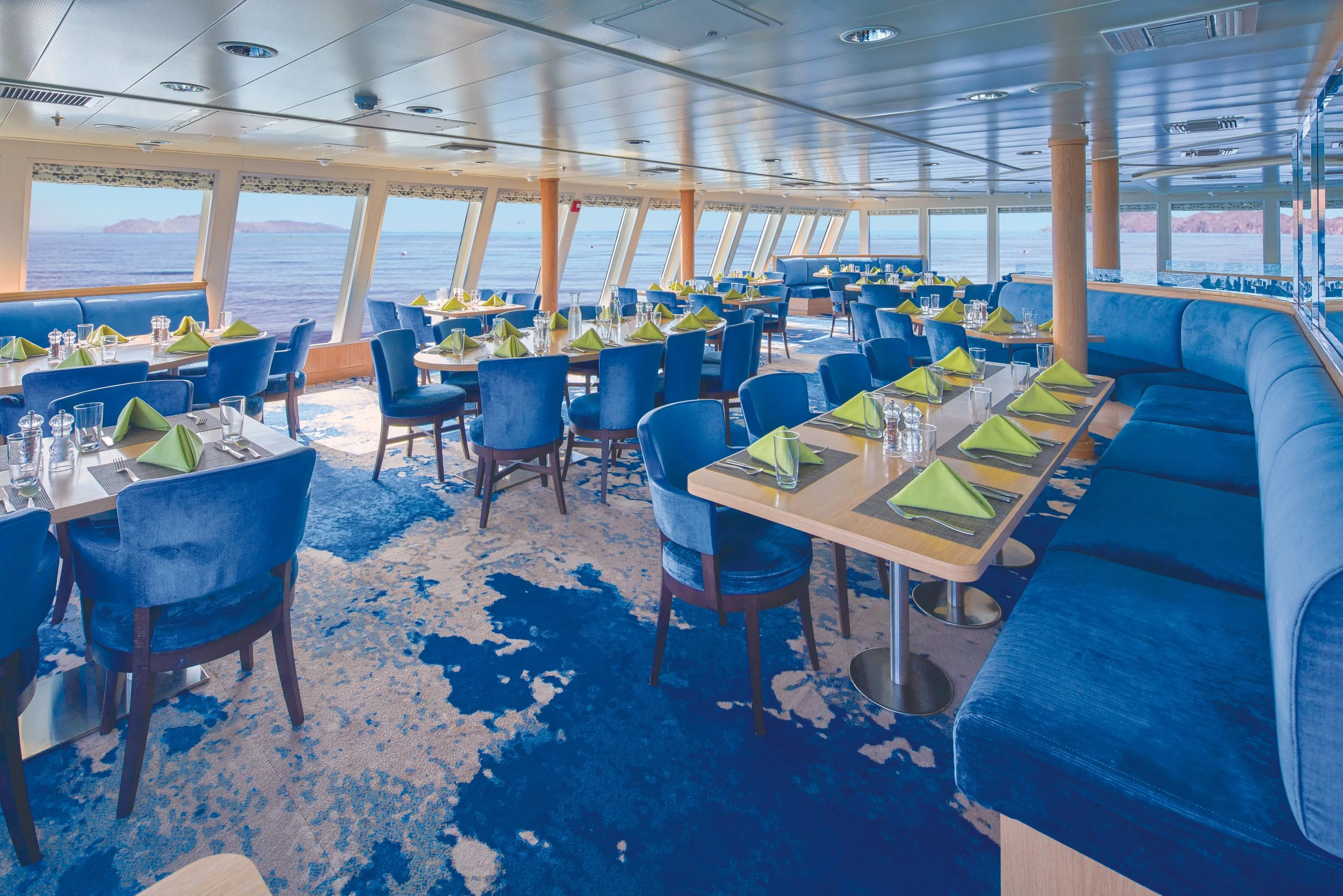 Dining area on a cruise ship with blue chairs, tables set with green napkins, and ocean view through large windows