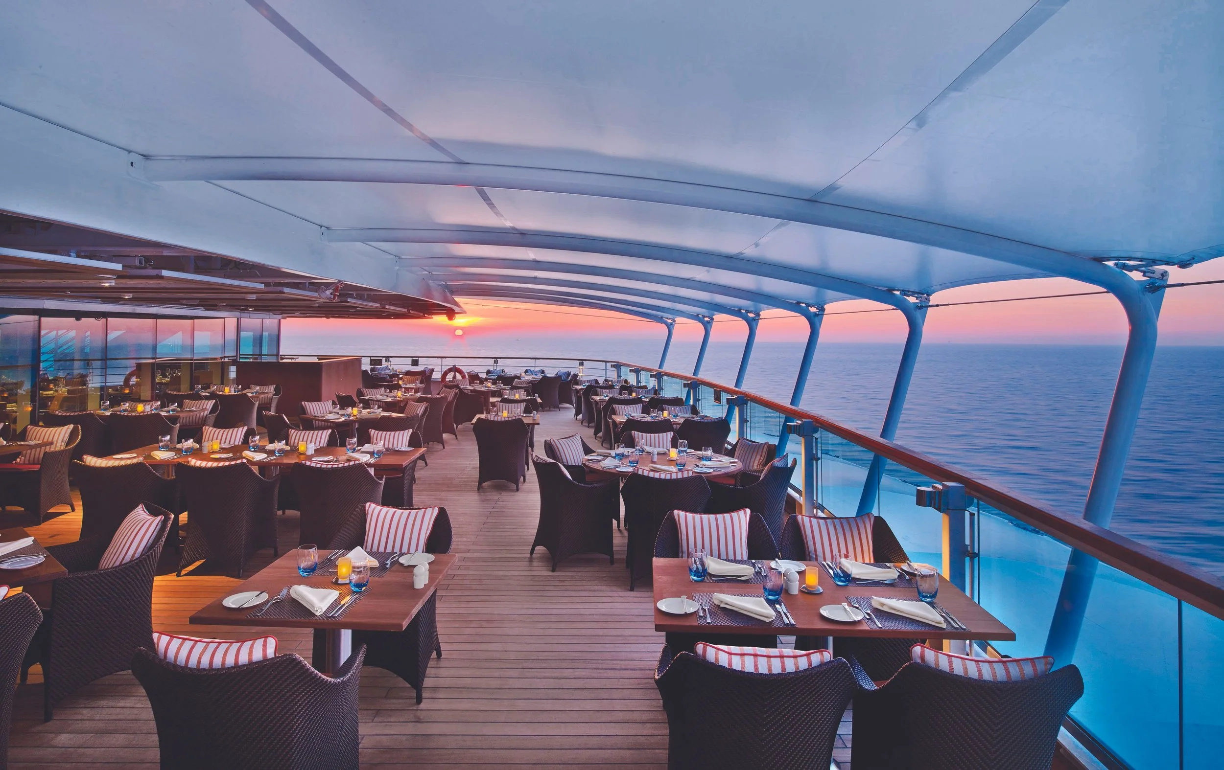 Empty outdoor dining area on a cruise ship deck at sunset with water in the background.