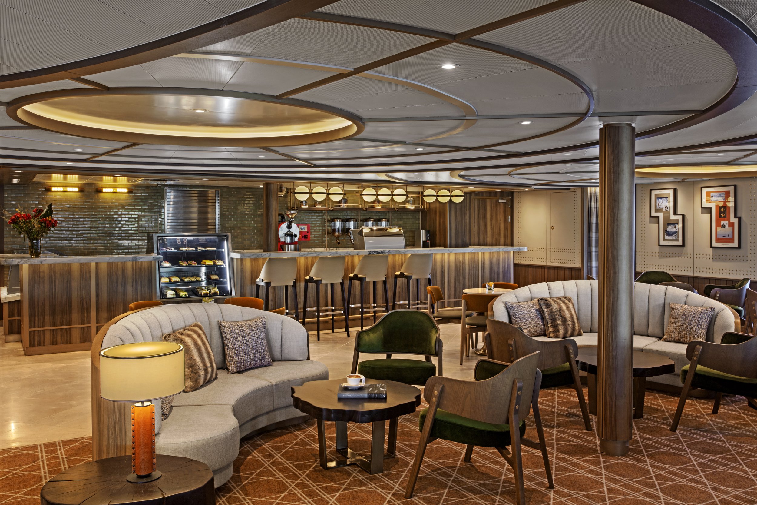 A cozy lounge area with curved sofas, wooden chairs, small tables, a lamp, and a bar with barstools in the background. The ceiling has modern circular design elements and warm lighting.