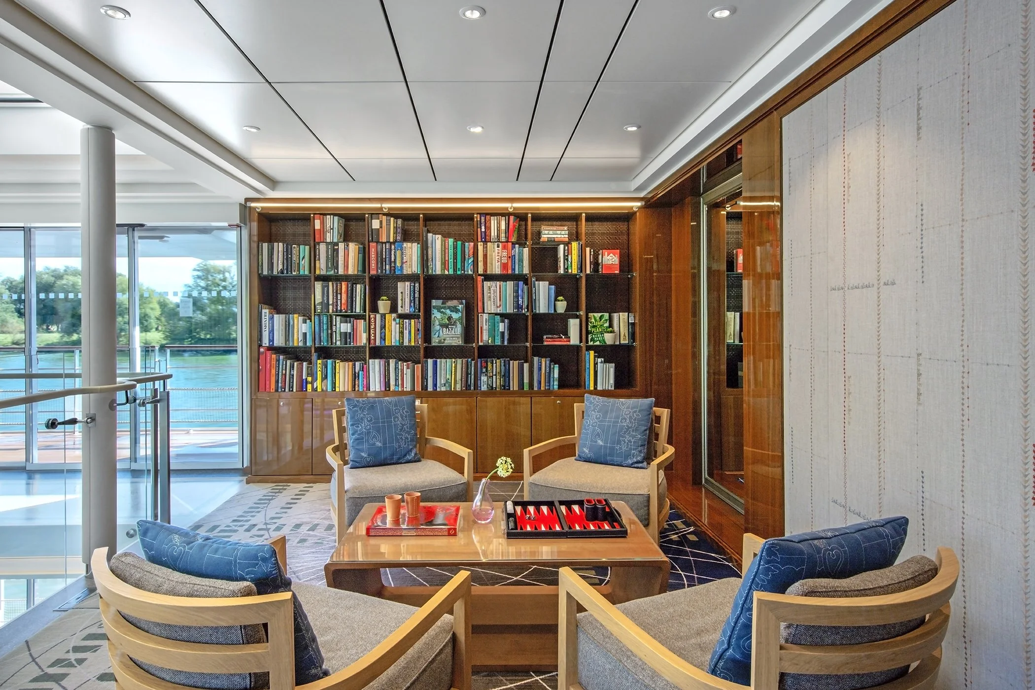 A cozy lounge area with four armchairs around a coffee table, a large wooden bookshelf filled with books, and a large window showcasing a view of a river and trees outside.