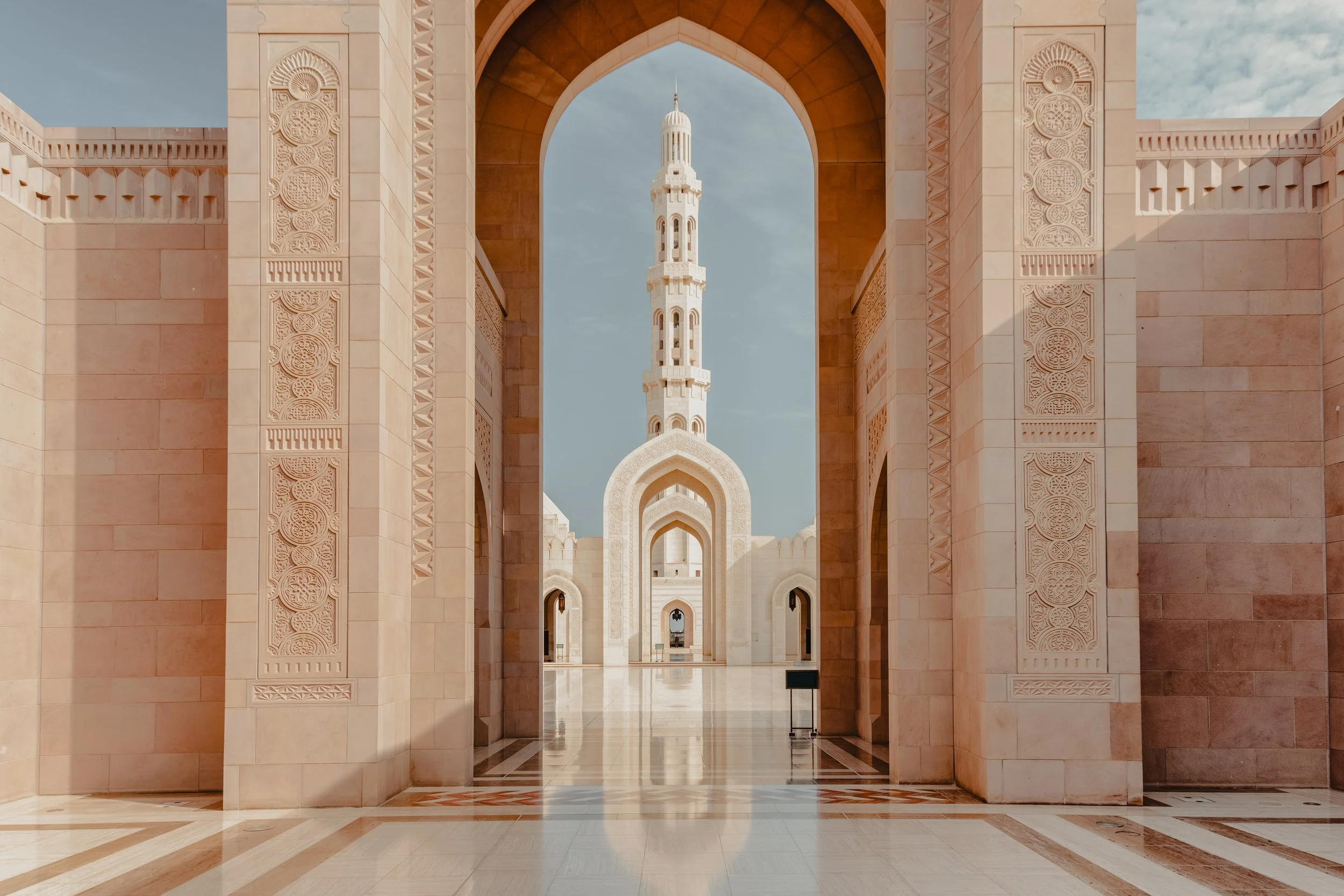 View of a mosque's interior with ornate pink and cream architecture, a tall minaret with a domed top in the background, under a partly cloudy sky.