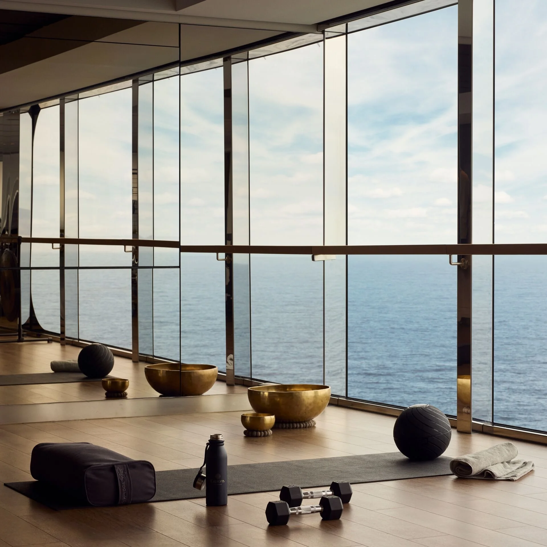 Interior of a yoga or fitness studio with large windows overlooking the ocean, containing yoga mats, weights, a water bottle, towels, exercise balls, and decorative bowls.