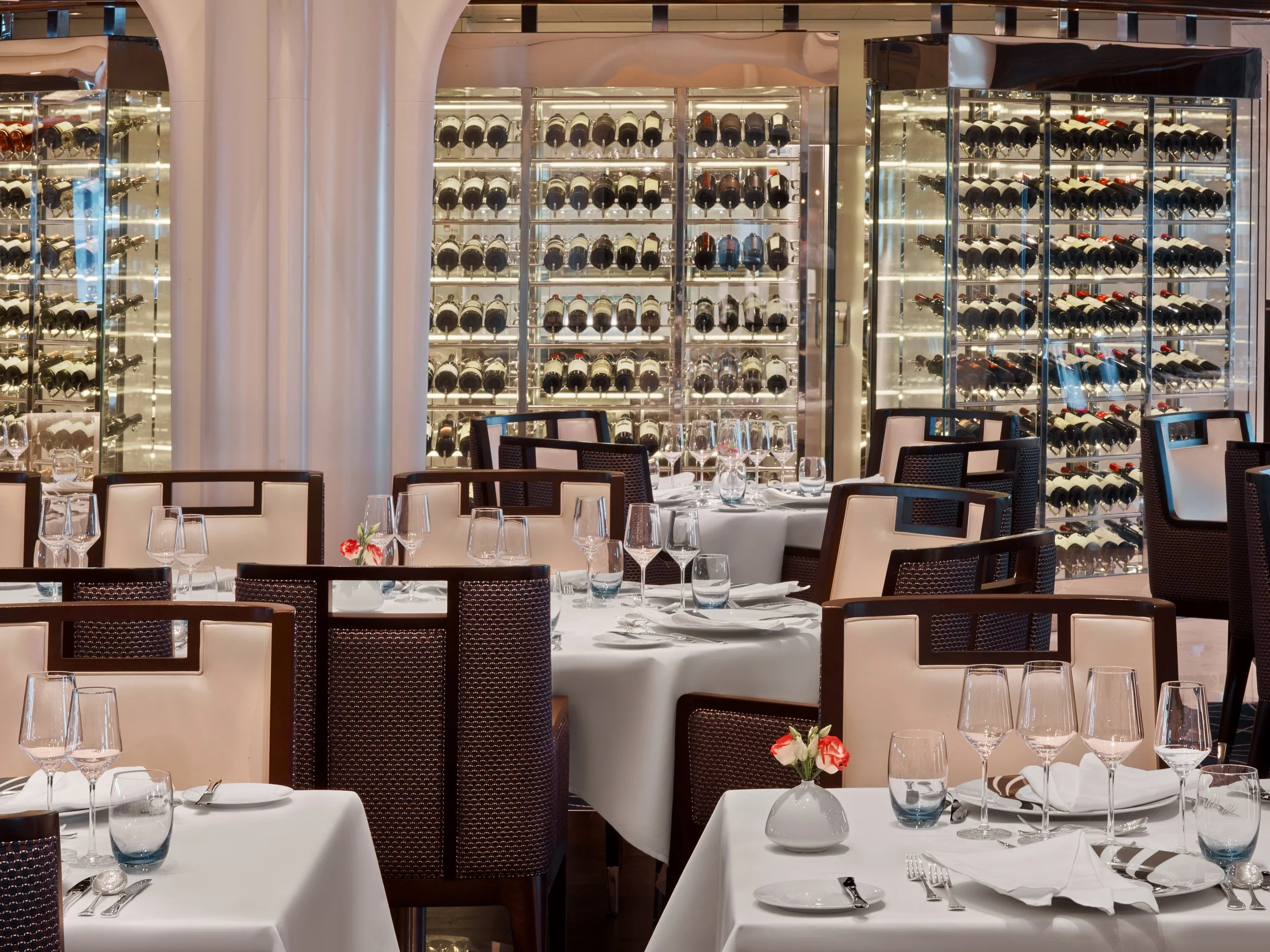 Upscale restaurant dining area with white tablecloths, empty wine glasses, and modern chairs, with wine bottles displayed on illuminated wine racks in the background.