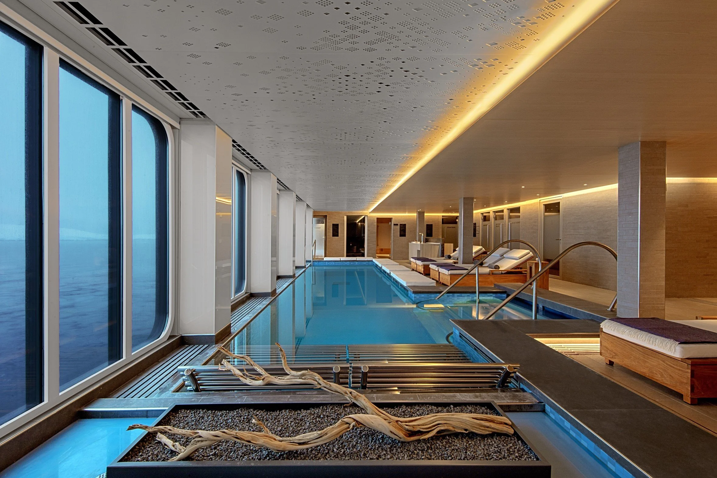 An indoor swimming pool area with large windows overlooking the ocean, modern lounge chairs, and ambient lighting.