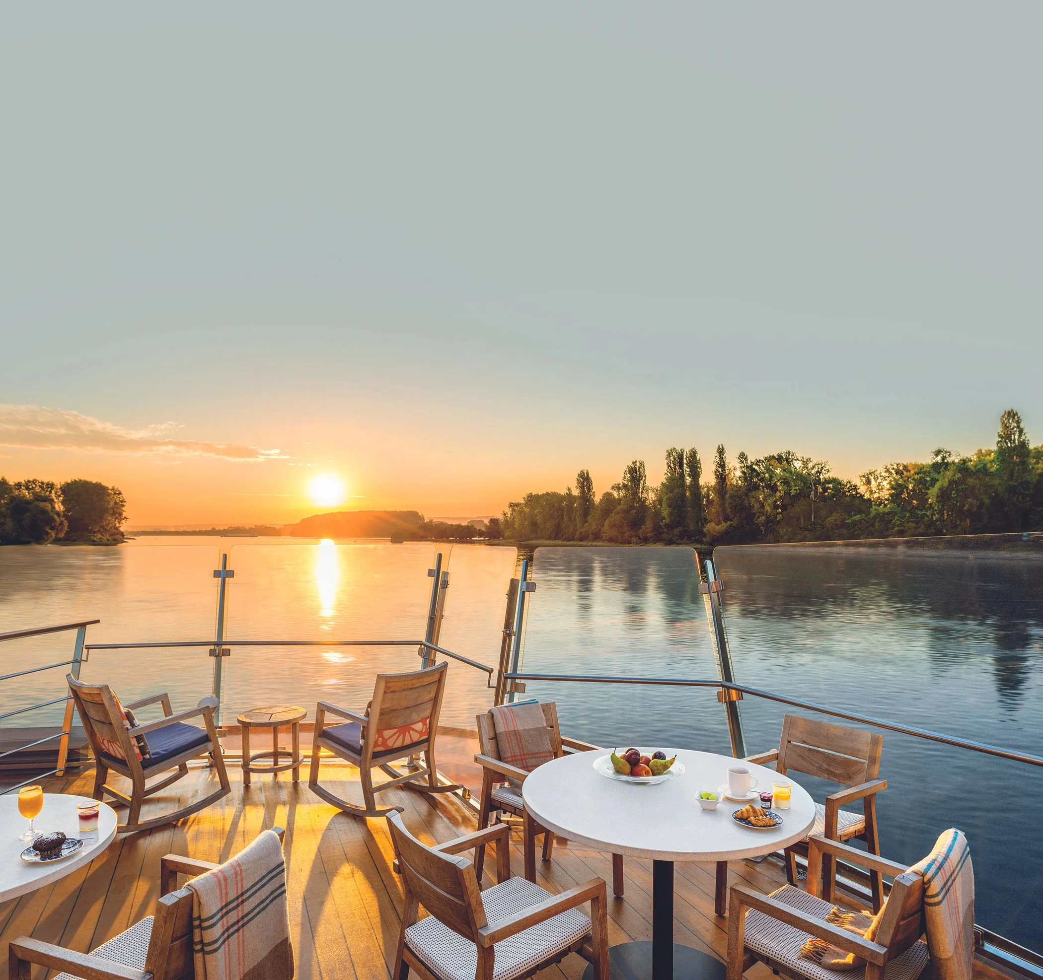 Sunset over a river viewed from a boat deck with chairs, tables, and breakfast spread including fruit, pastries, coffee, and juice.