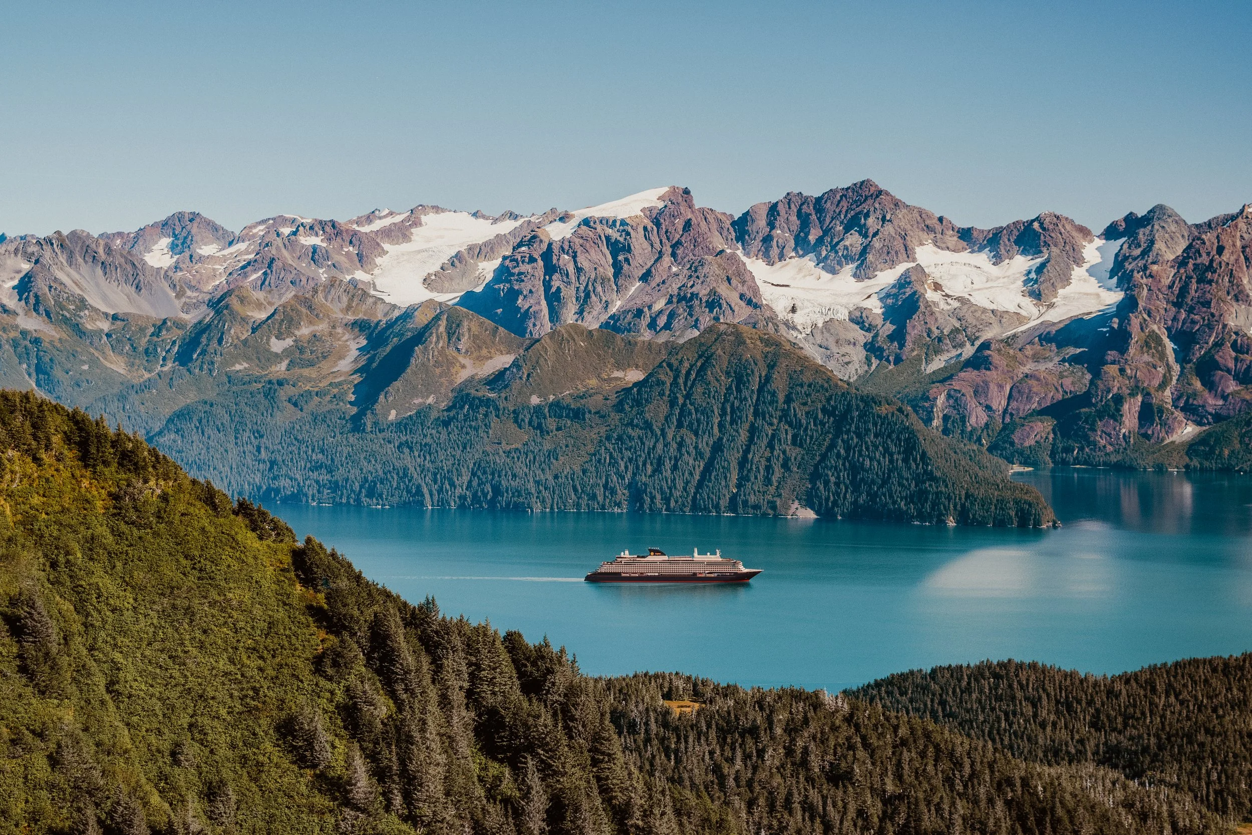 A cruise ship sailing on a blue lake surrounded by green forests and snow-capped mountains.