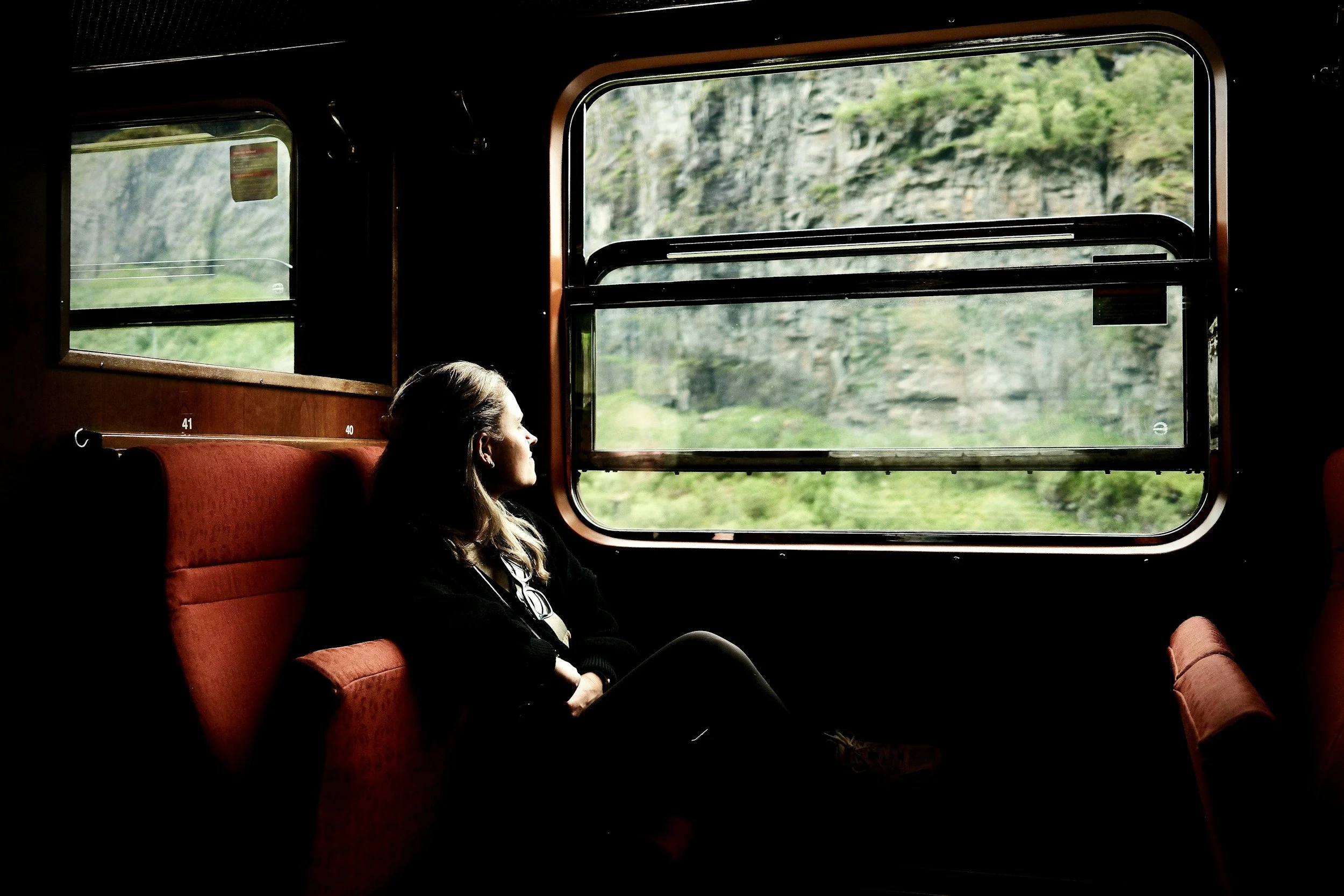 A woman sitting on a train seat, looking out the window at green hills and rocky cliffs outside.